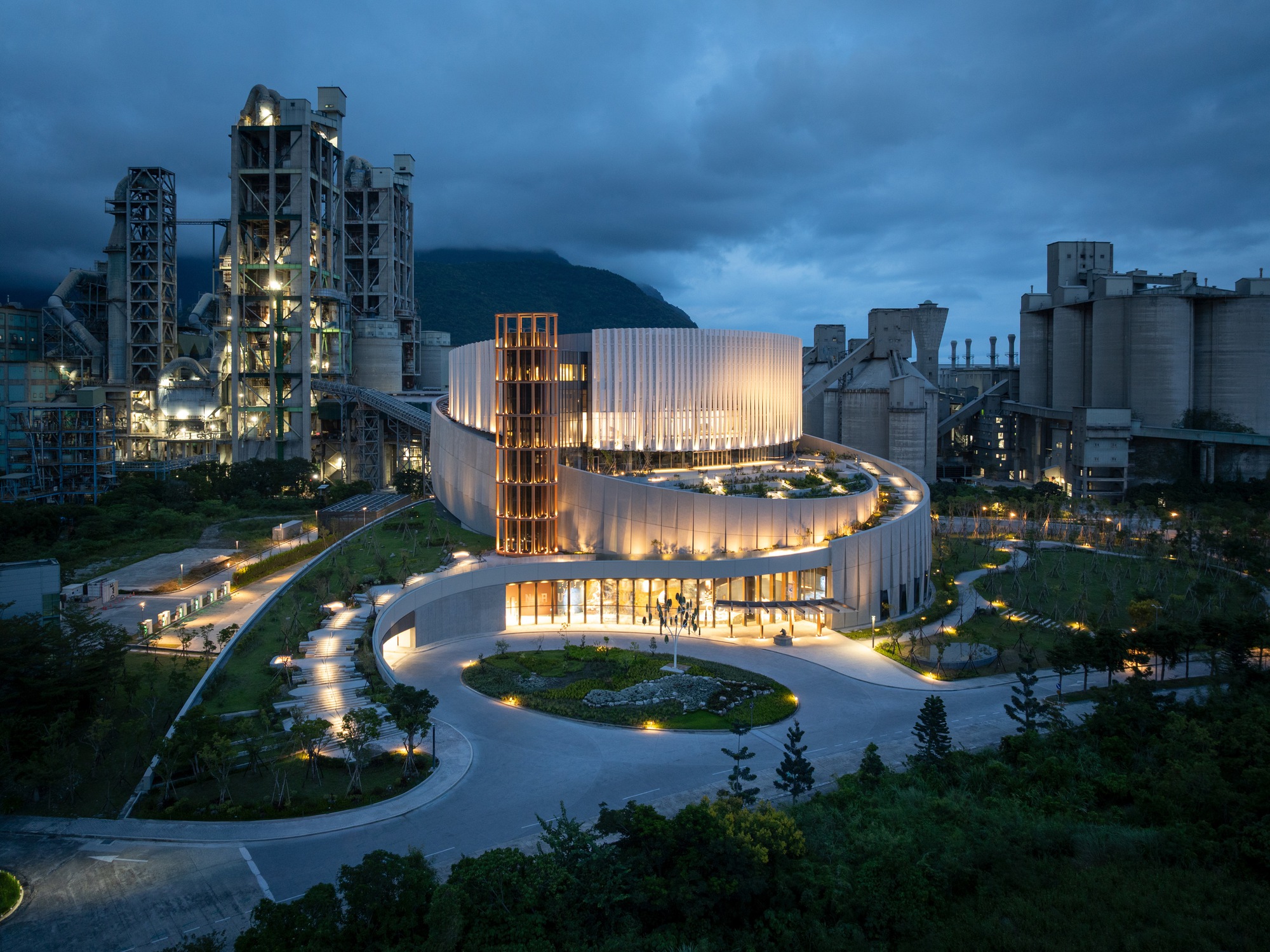Elevated view of the circular building ringed by lit paths and a landscaped island, with cement plant towers under heavy dark clouds