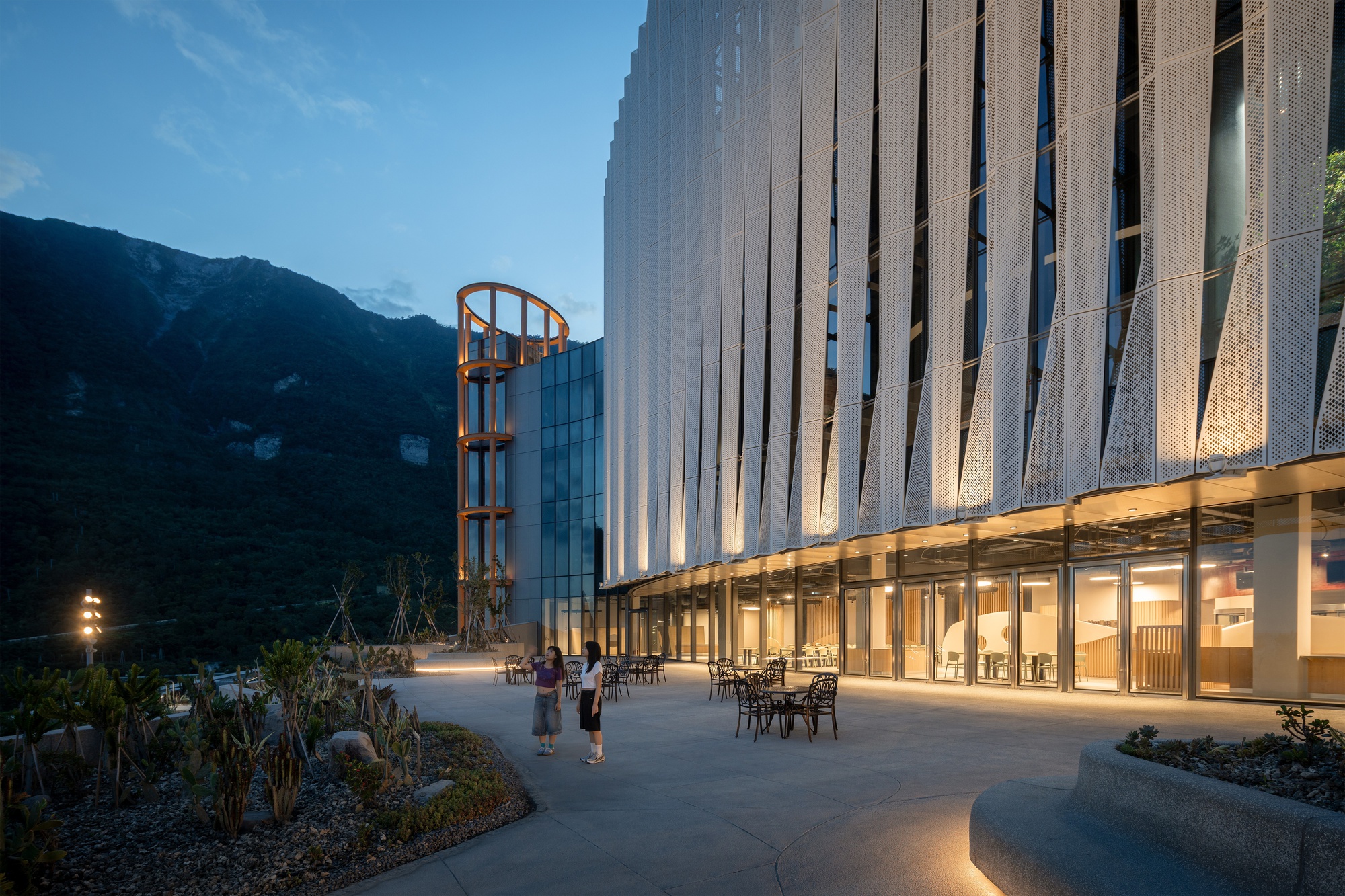 Terrace with café tables and two people in front of the pleated perforated metal facade, a mountain rising behind at dusk