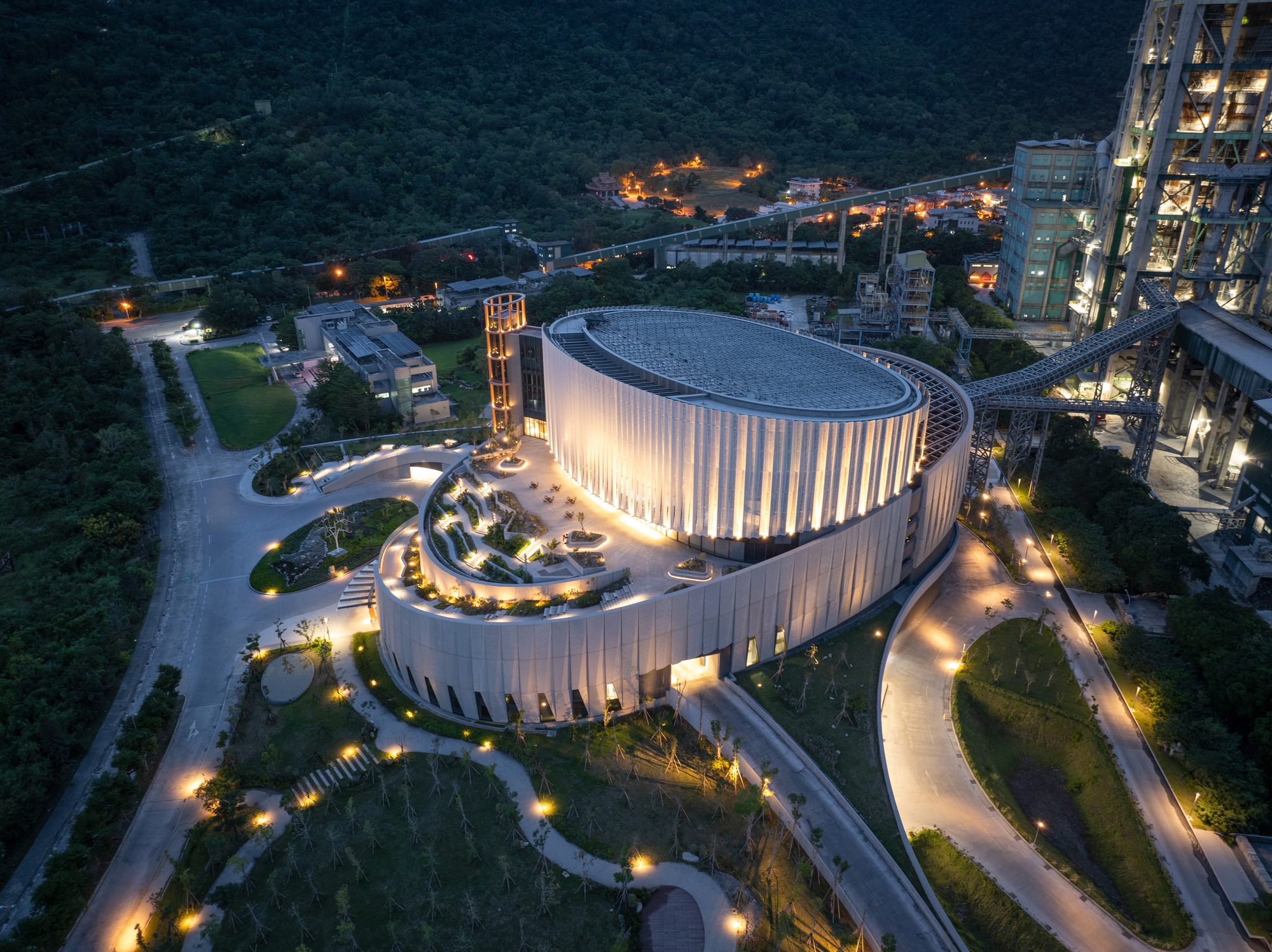 Aerial view of the oval building with its lit rooftop garden and pleated facade, surrounded by curving paths and forest at night