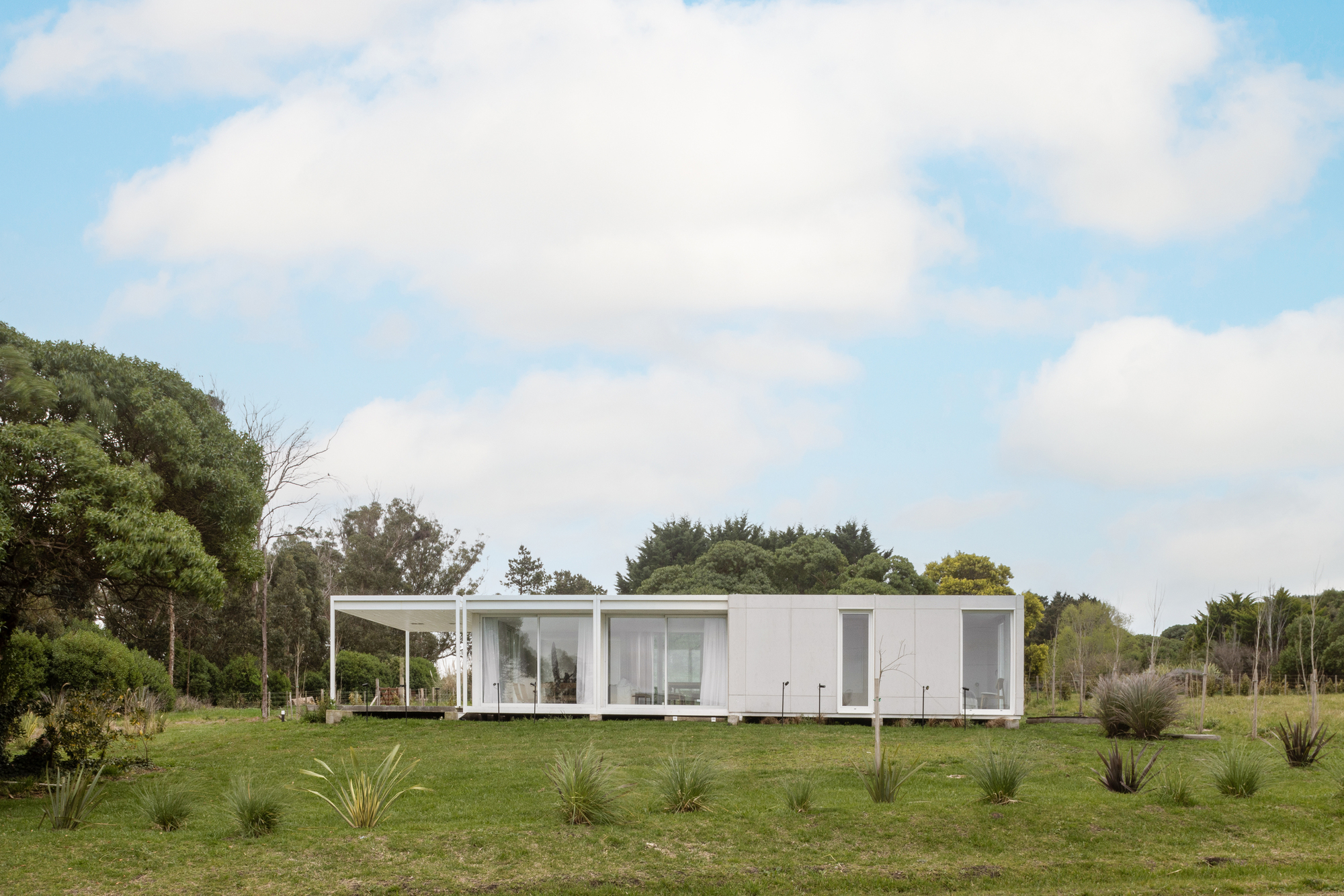Single-storey white panel house with a glazed porch end, set in a grass meadow with spiky plants under scattered clouds
