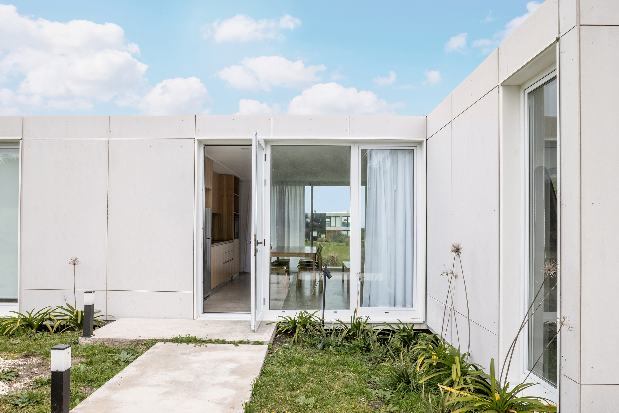 Open entry door in a white panel wall, with a concrete path crossing a planted lawn toward the kitchen