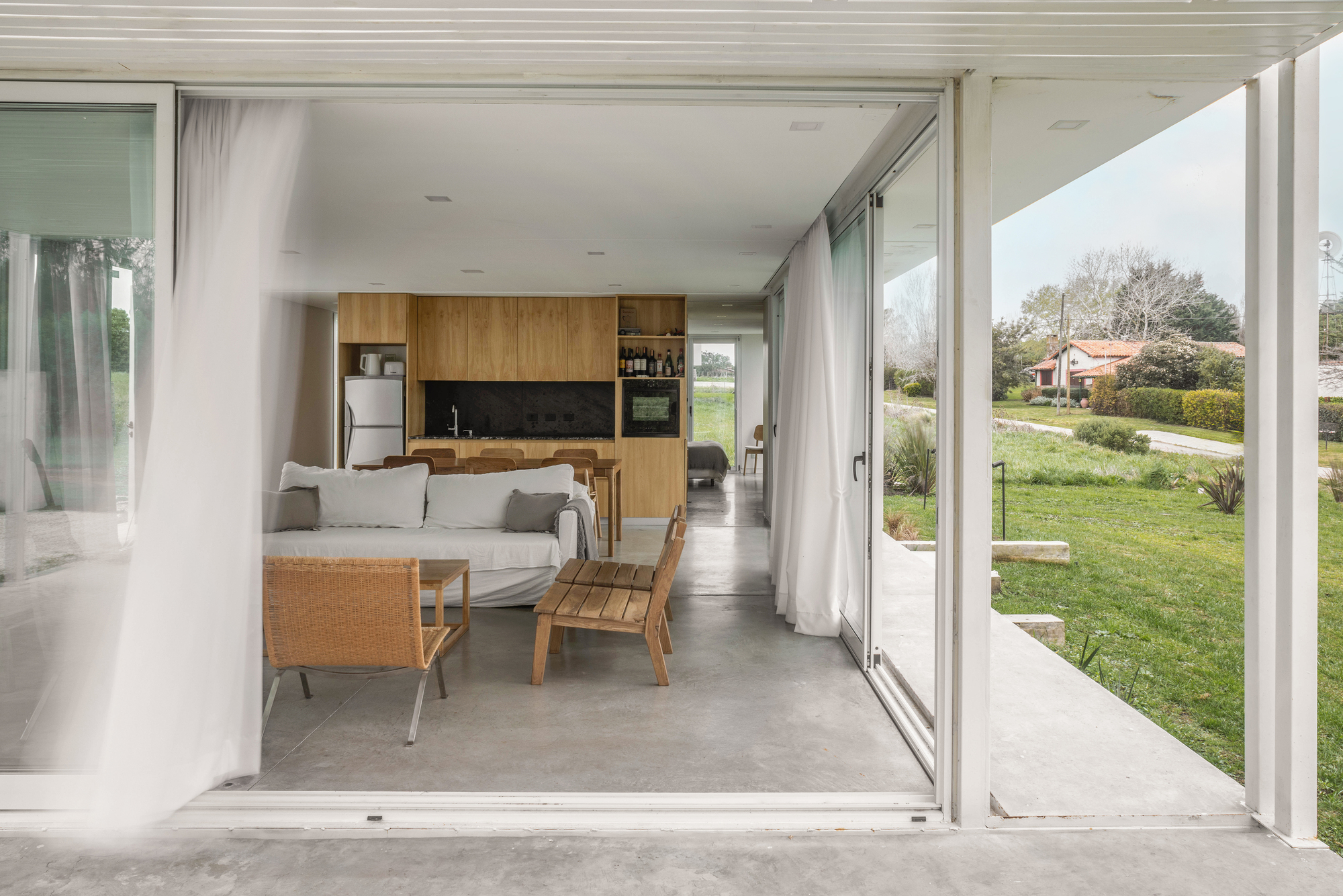 Living area seen through sliding glass and sheer curtains, with wicker chairs, a timber kitchen block and a concrete floor