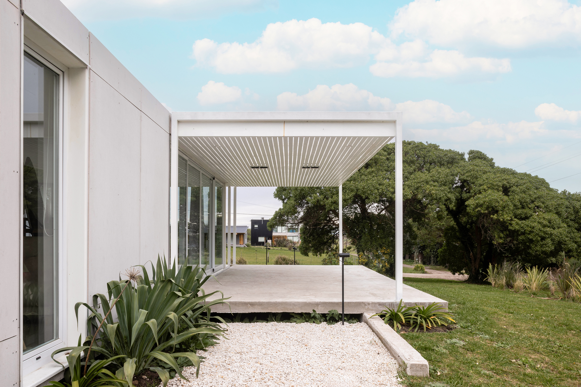 Slatted white canopy over a concrete terrace with glazed doors, beside a gravel bed of agave-like plants in daylight