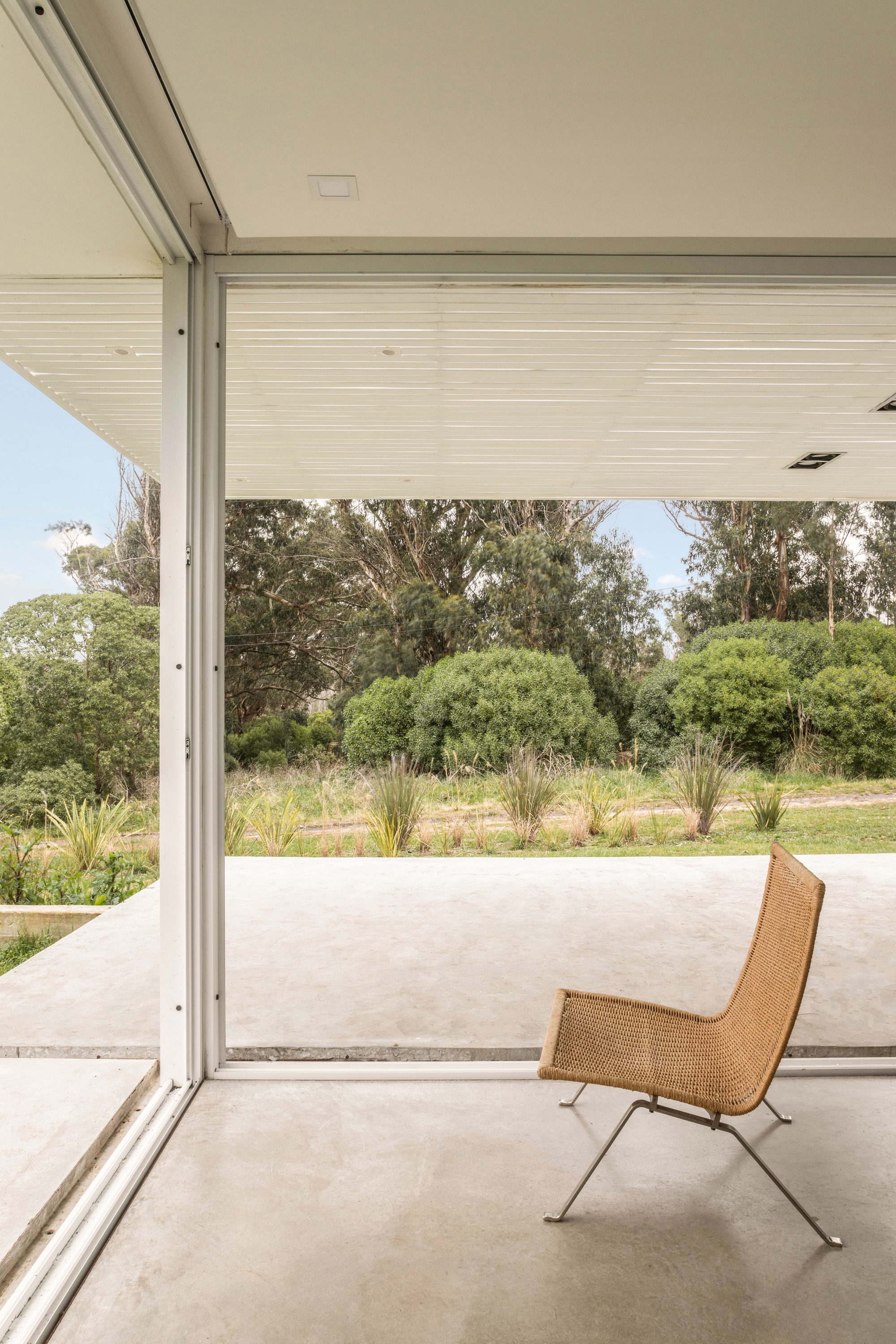 Woven wicker lounge chair on a polished concrete floor, framed by white mullions and a row of shrubs beyond