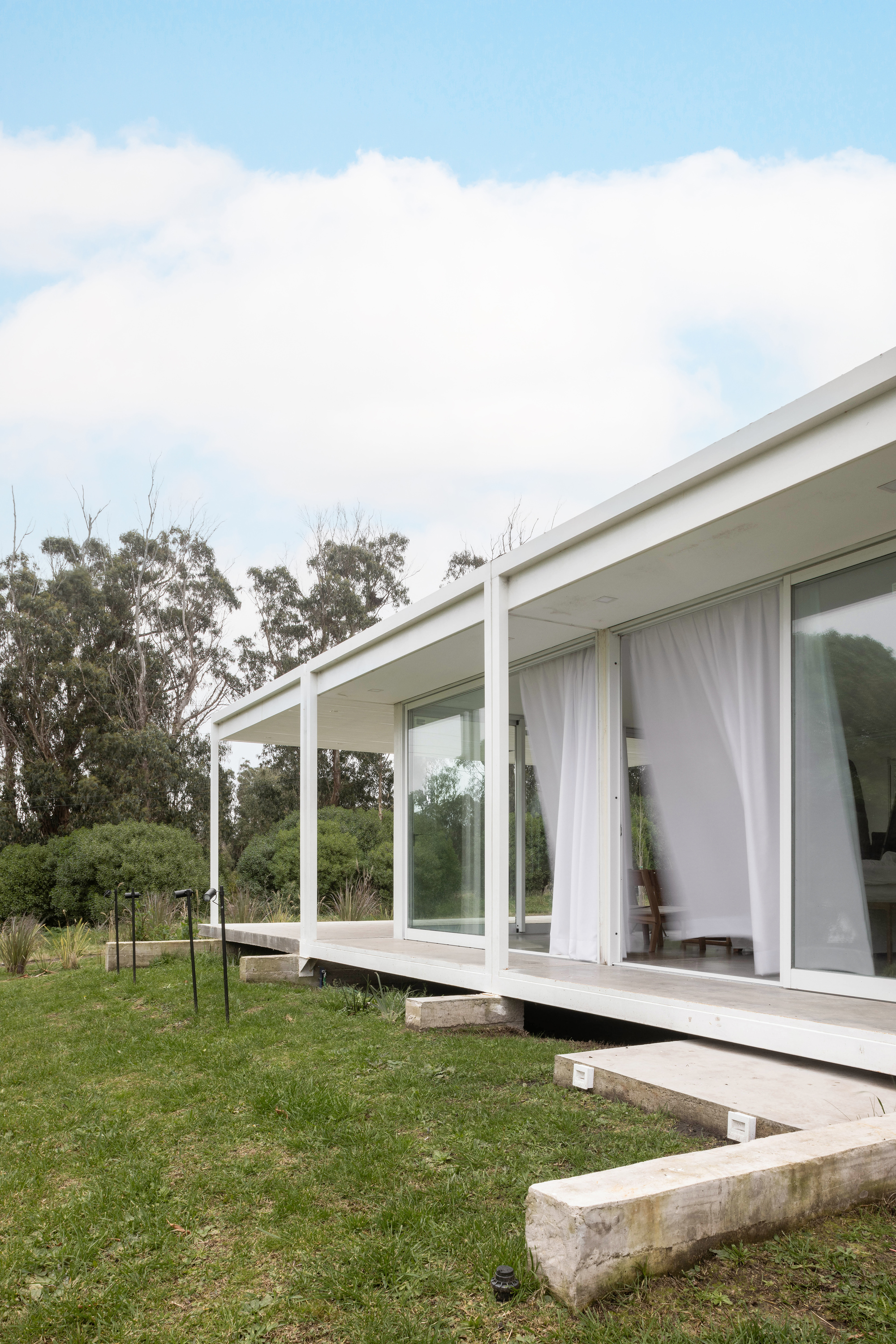 Corner of the raised white porch with slim columns and billowing curtains, with timber step blocks set in the lawn