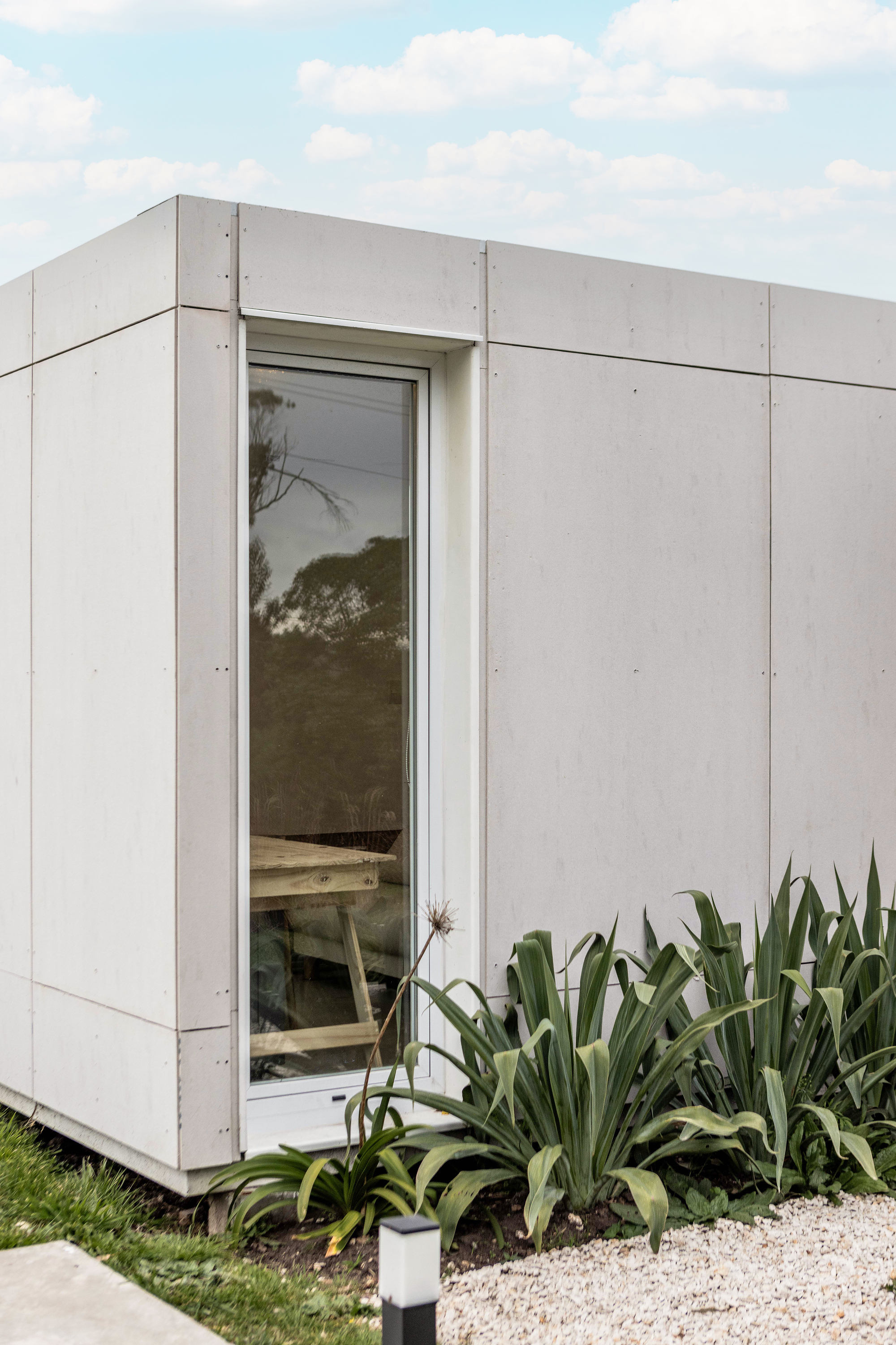 Corner of the grey cement panel facade with a tall narrow window and agave plants above white gravel