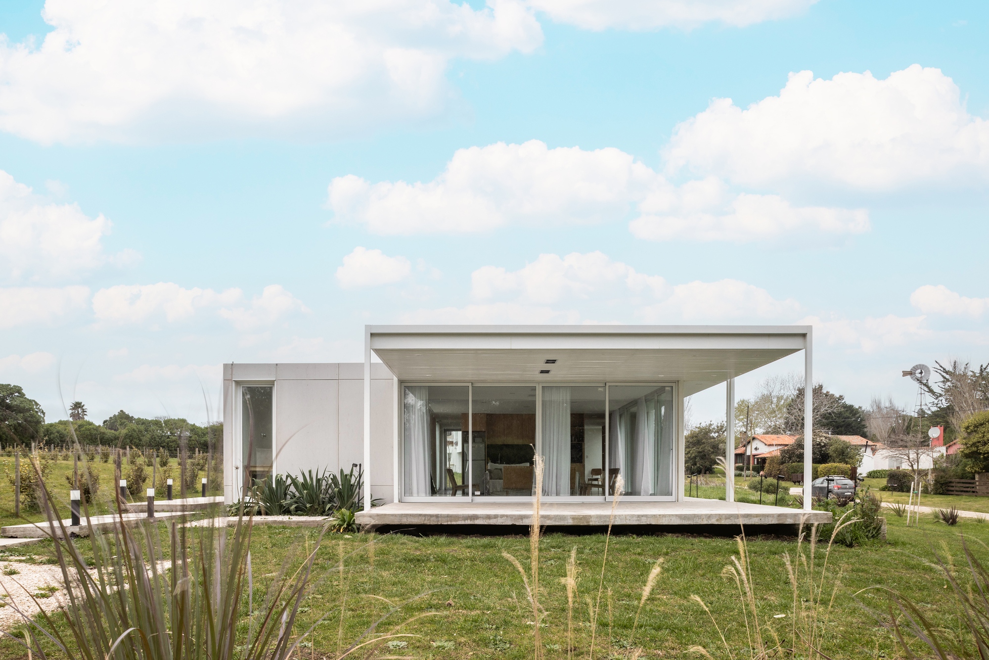 Front view of the glazed living pavilion on a concrete platform, framed by tall grasses under a cloudy blue sky