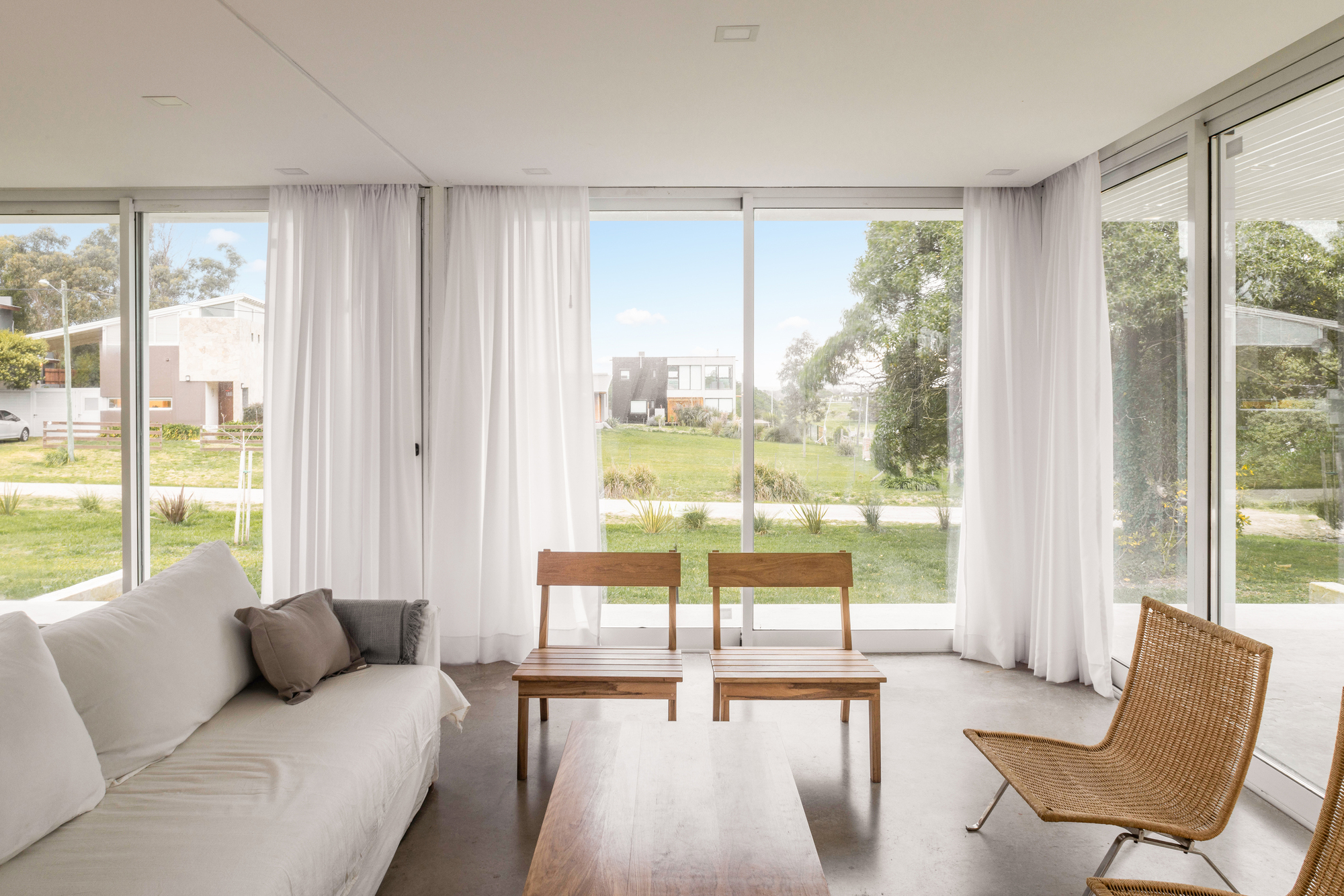 Living room corner with sheer white curtains, timber chairs and a low table facing floor-to-ceiling glass and a green lawn