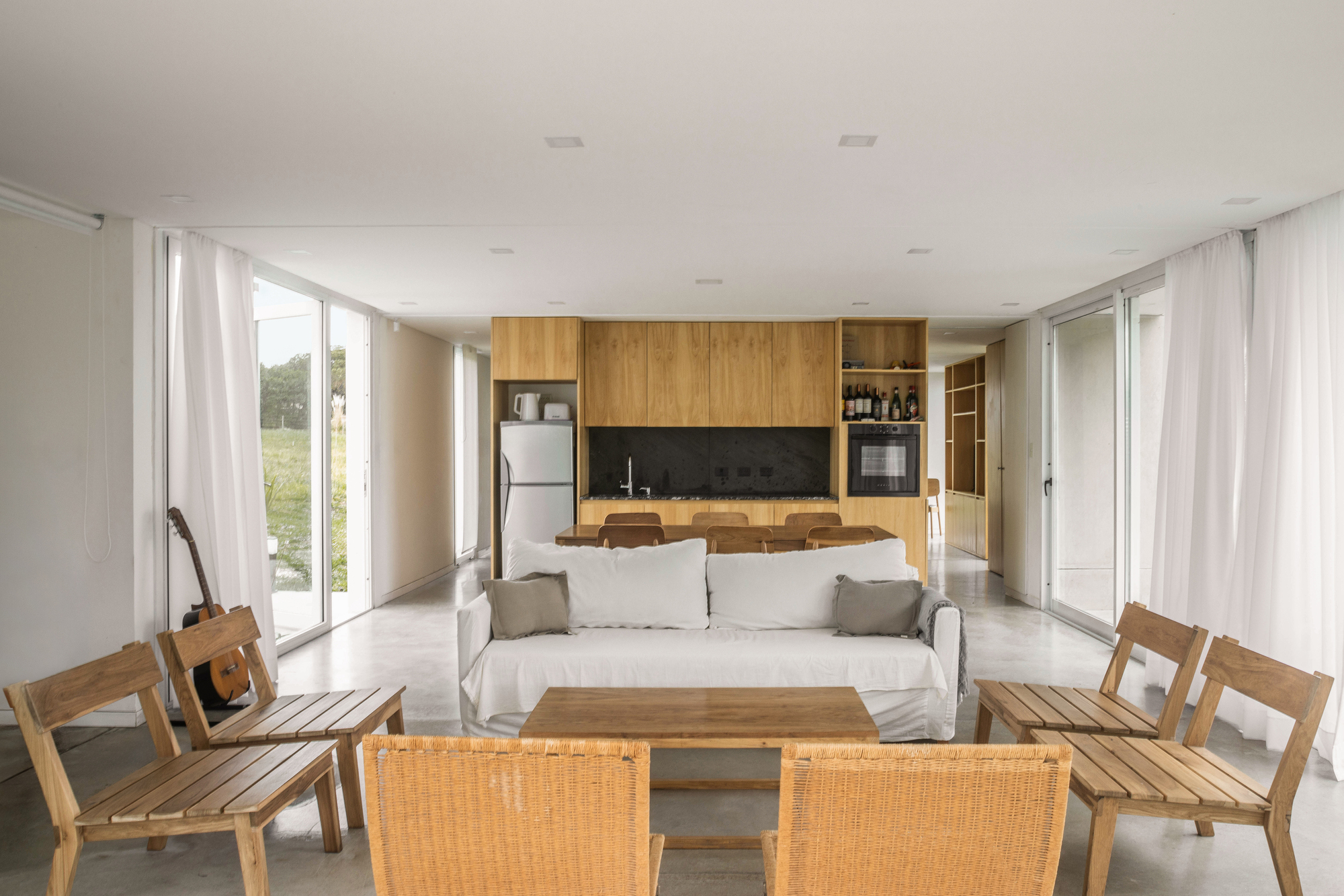 Open living and kitchen space with oak cabinetry, white sofa, wicker and slatted timber chairs, and a guitar by the window