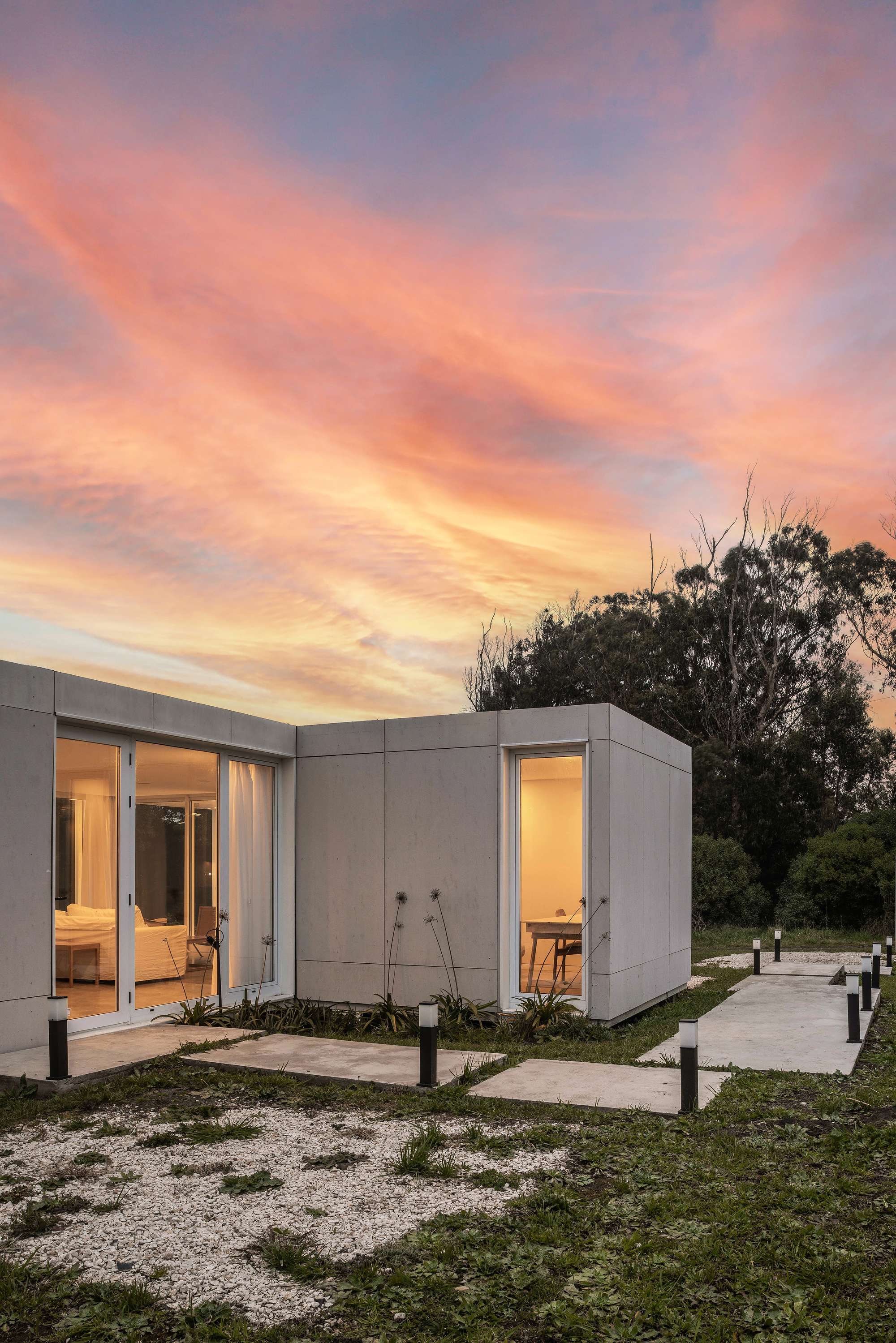 Lit panel-clad volume with glazed corners and stepping-stone paths with bollard lights under a pink and orange dusk sky
