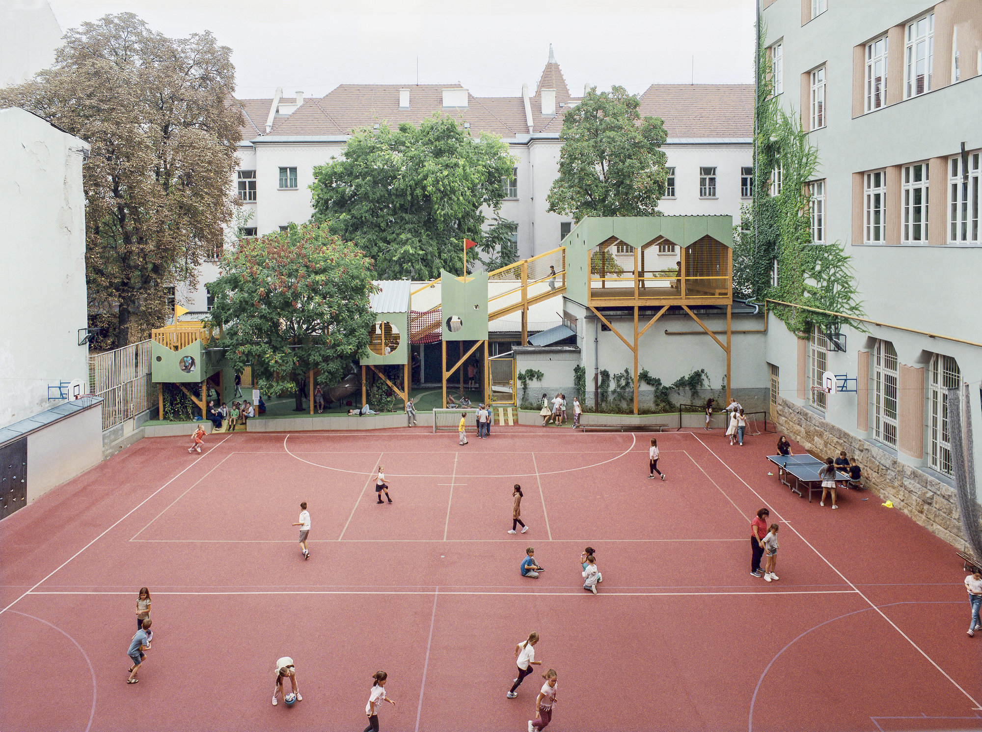 High view of a red sports court with children playing below green play structures on timber stilts between courtyard buildings