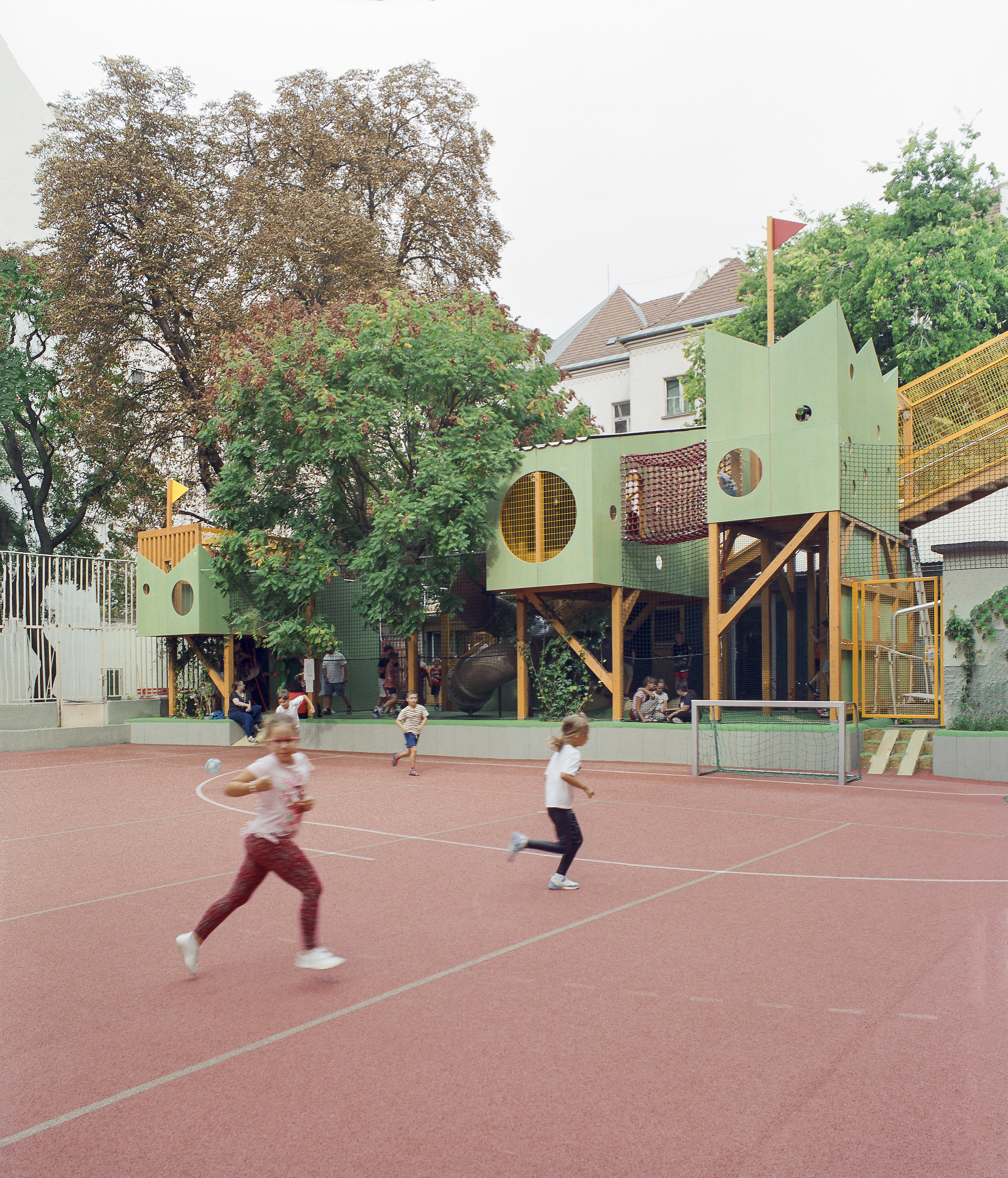 Children running across the red court in front of a green play pavilion with round windows, netting and a small goal