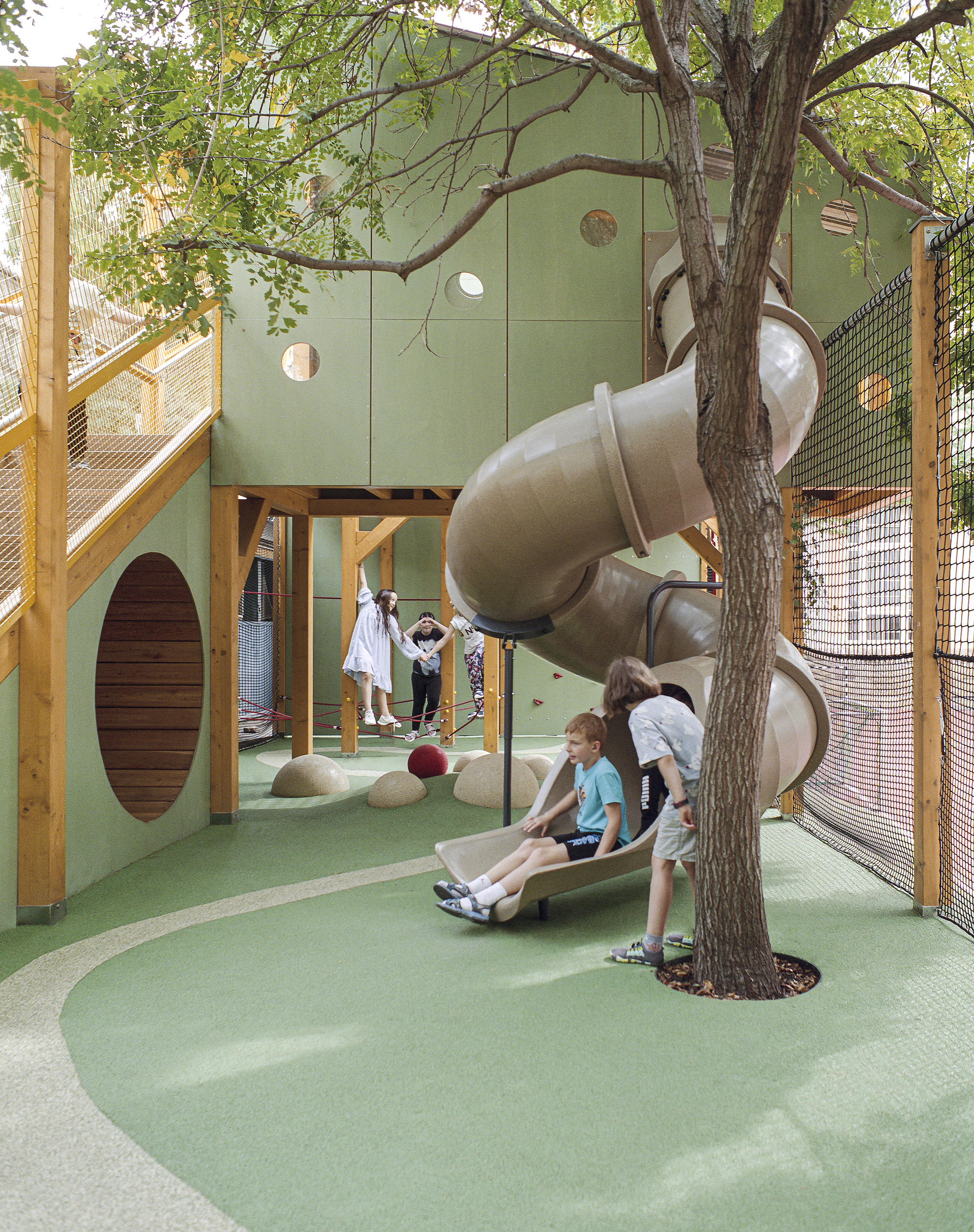 Spiral tube slide beside a tree trunk on green rubber flooring, with children sitting at its base and a round timber panel