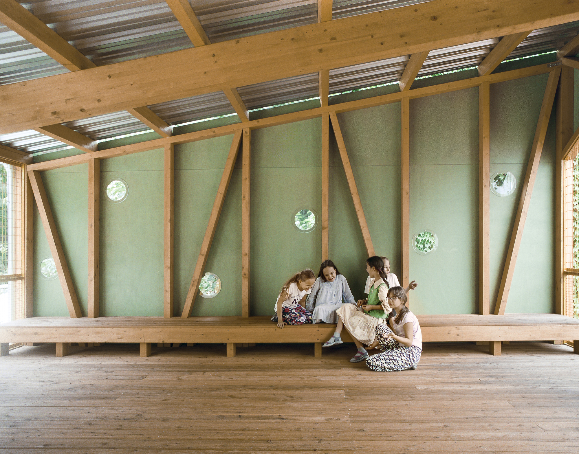 Children seated on a long timber bench inside a pavilion with green panel walls, diagonal braces and round porthole windows