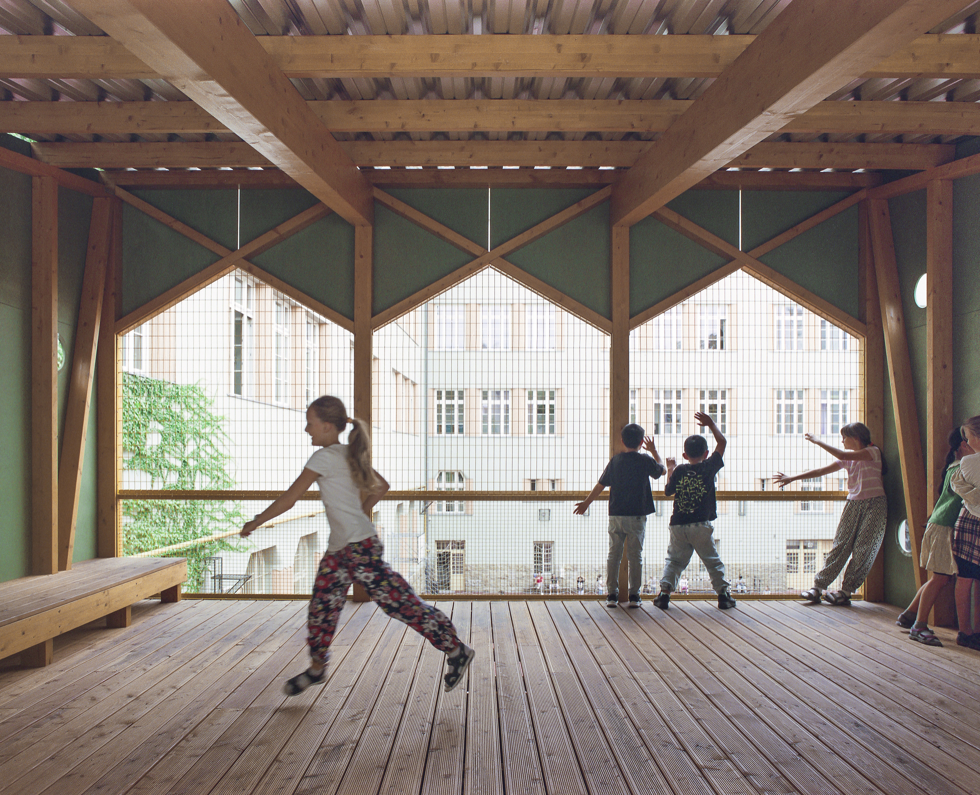 Girl running across a plank floor while other children stretch at a mesh opening framed by timber, facing the courtyard facades