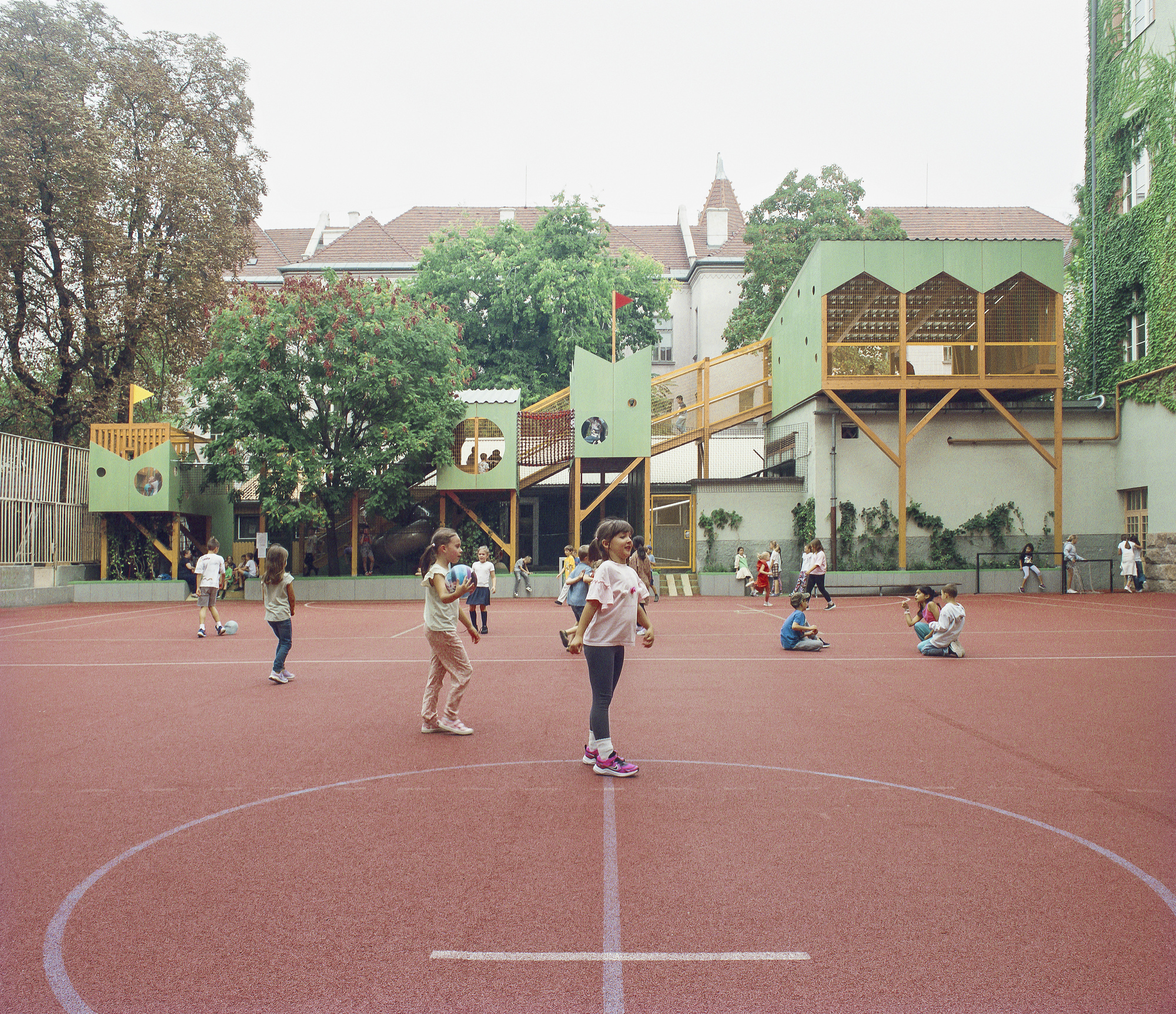 Children with a ball at the court's center circle, with the green stilted play structures and ivy-covered wall behind under overcast sky