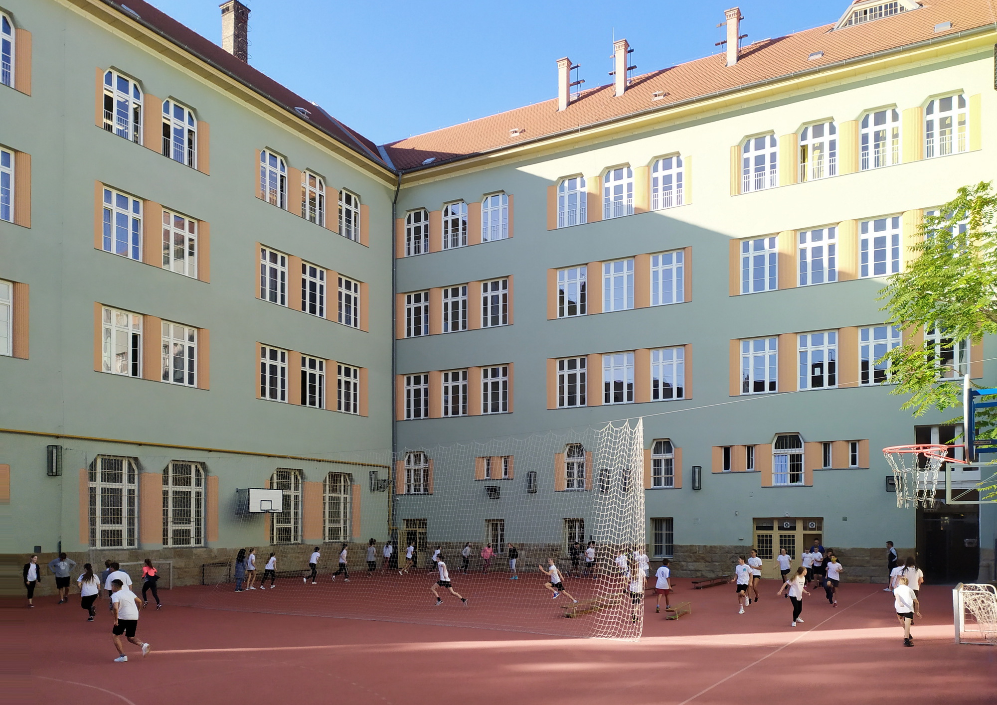 Schoolyard courtyard framed by pale green facades with orange window surrounds, children playing on the red court in strong sunlight