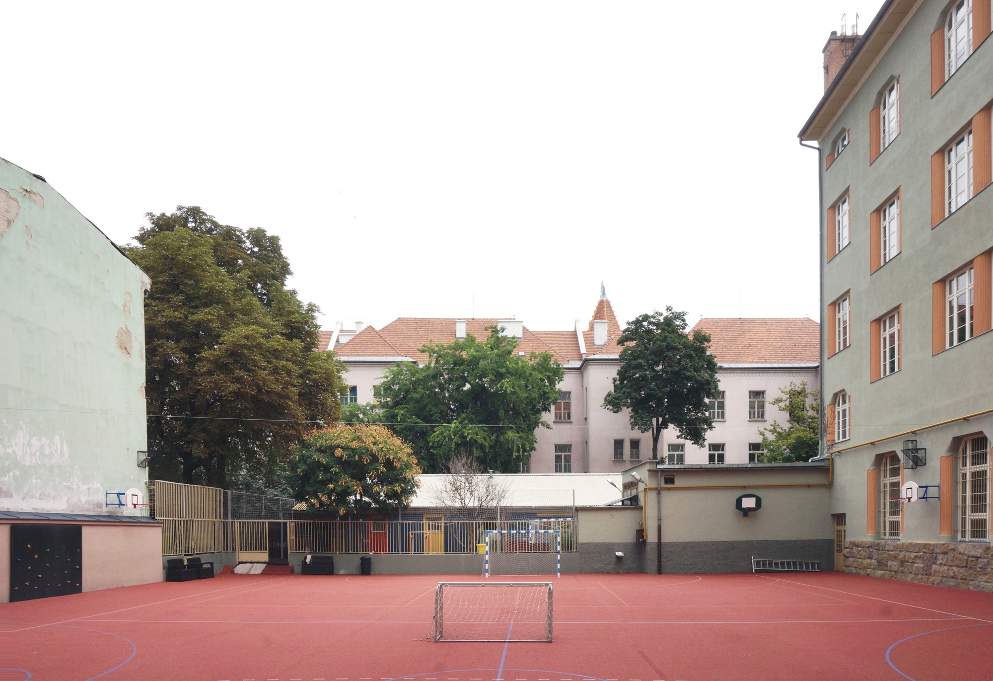 Red sports court with a small goal, enclosed by a green facade, a mesh fence and leafy trees under overcast sky