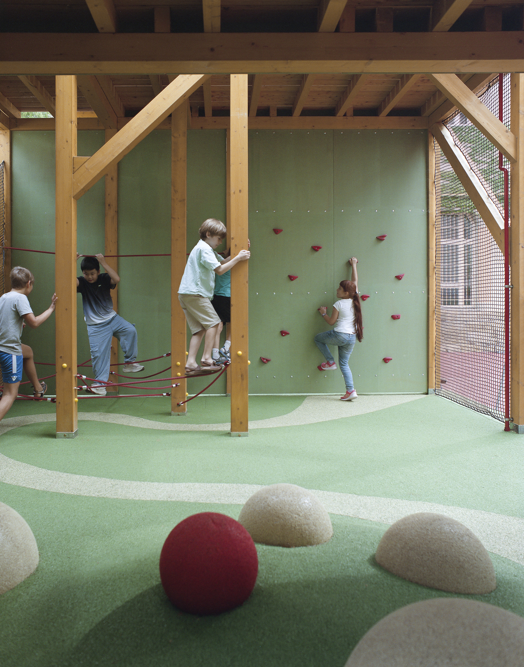Timber-framed play pavilion with a green climbing wall, rope net and rubber hills, children climbing in soft daylight