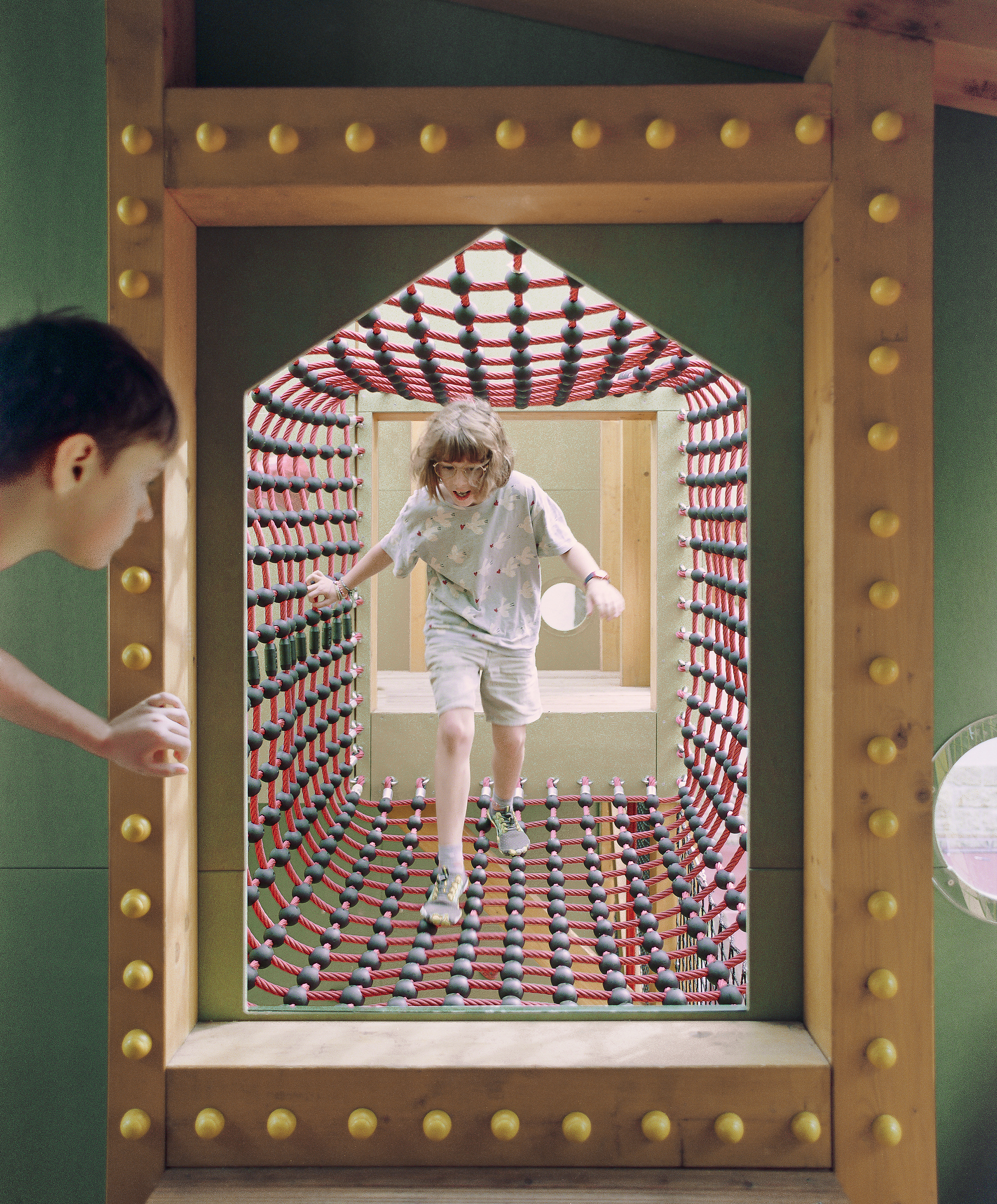House-shaped opening studded with wooden knobs, framing a child crossing a red rope net tunnel while another peers in