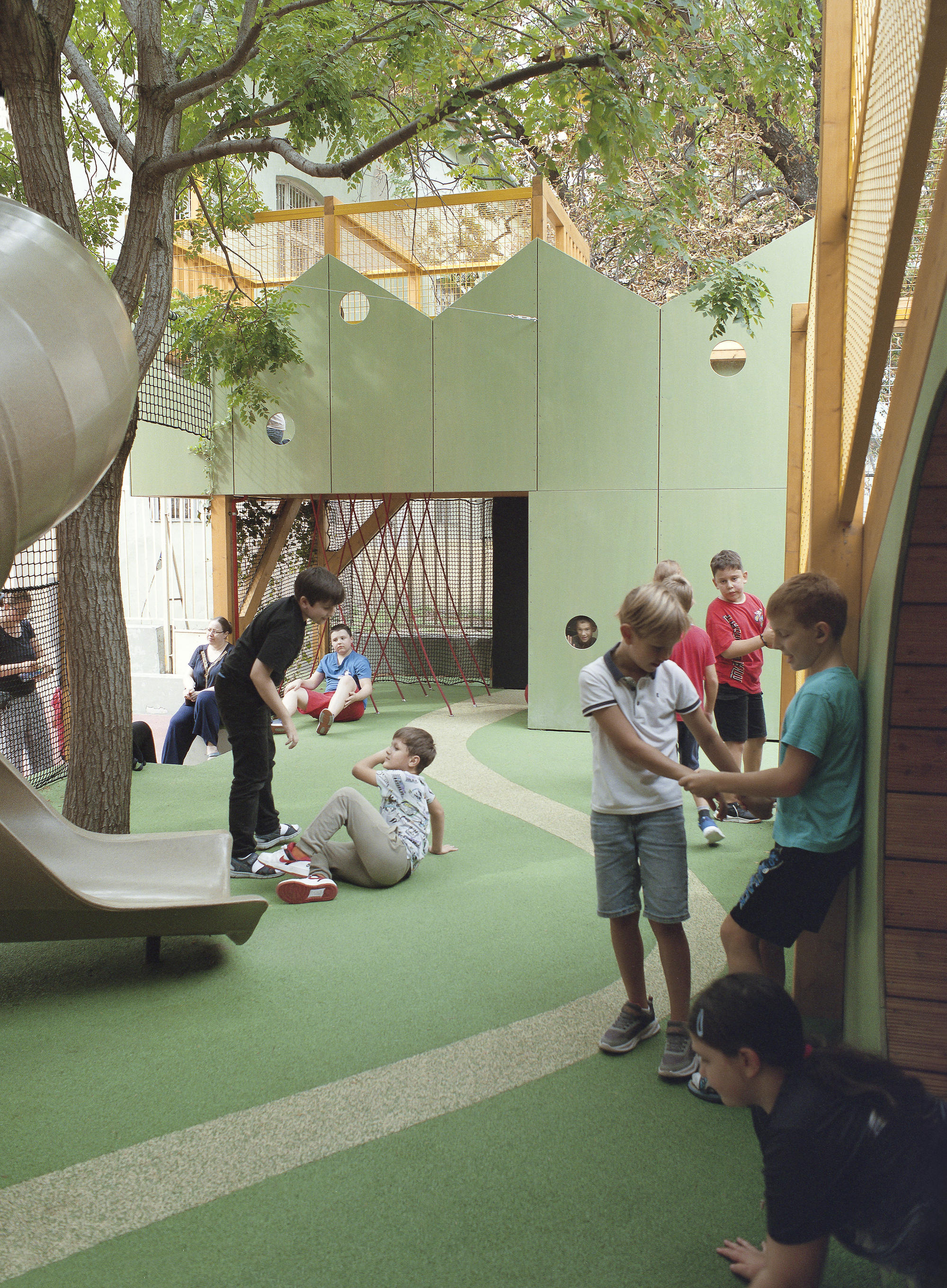 Green playground with a gabled panel wall, slide and rope structure, children gathered beneath a leafy tree in dappled light