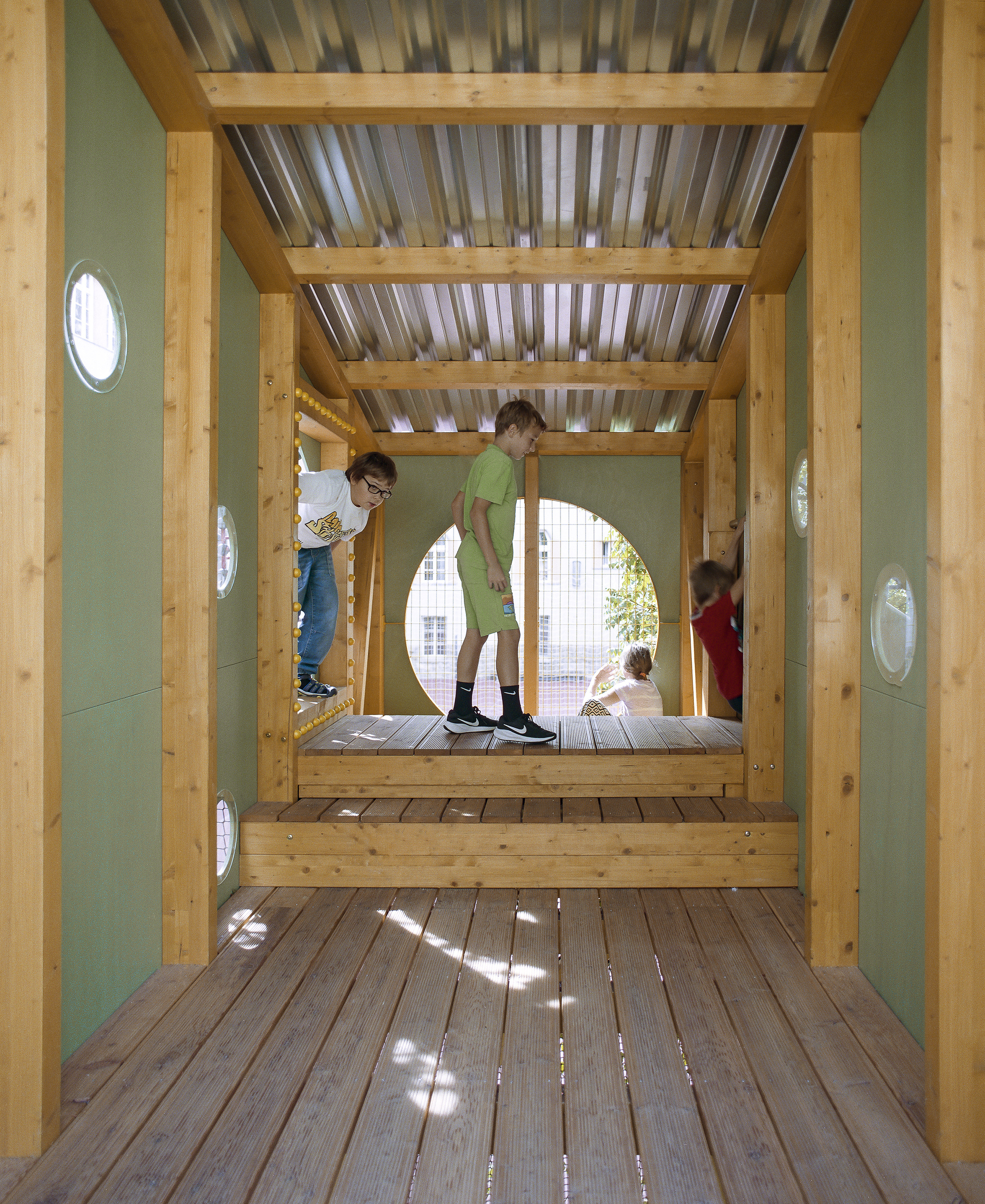 Timber-framed passage with green panel walls, round porthole windows and corrugated roof, children climbing a step platform in dappled daylight