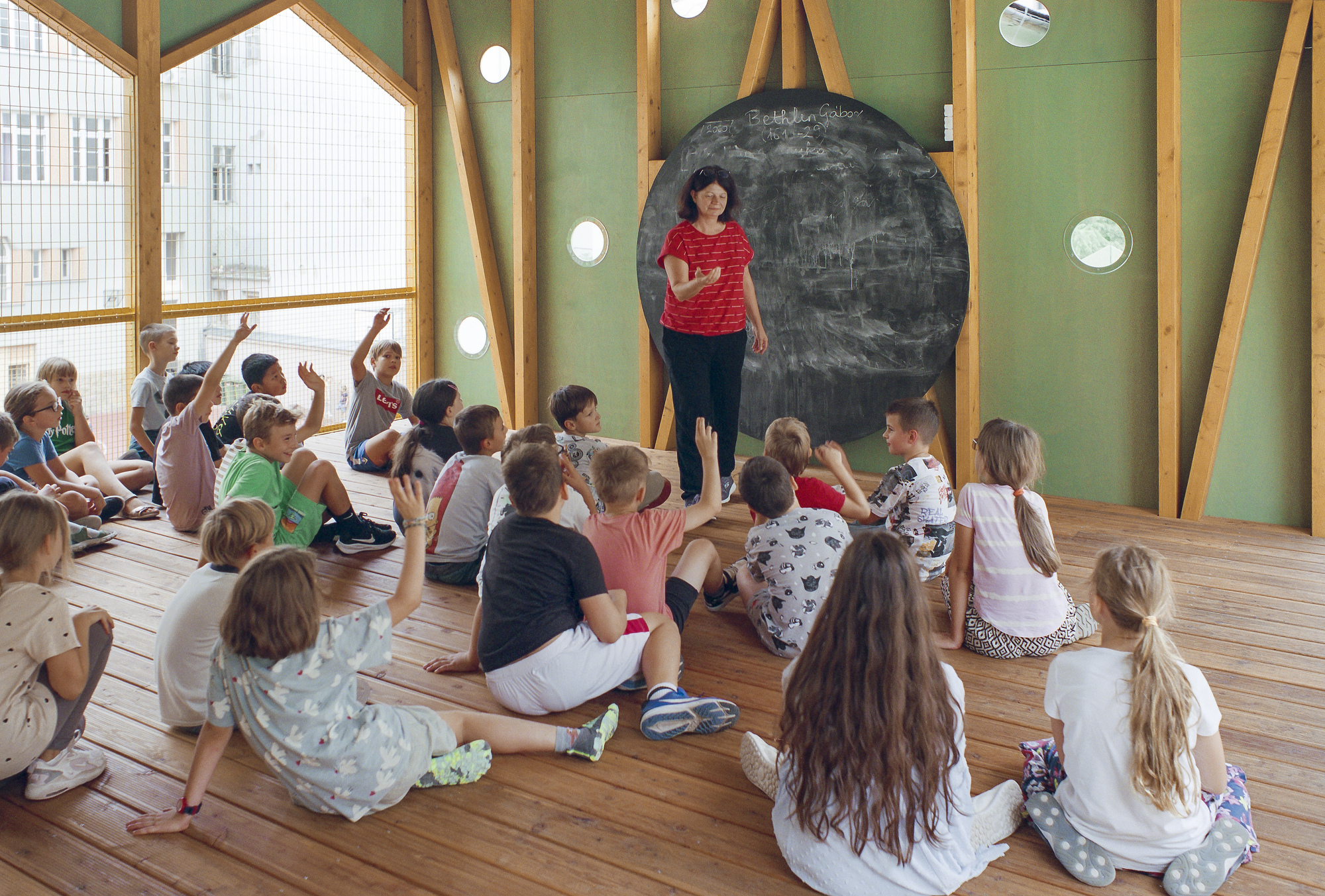 Children seated on a plank floor facing a teacher beside a round chalkboard, with green walls and porthole windows