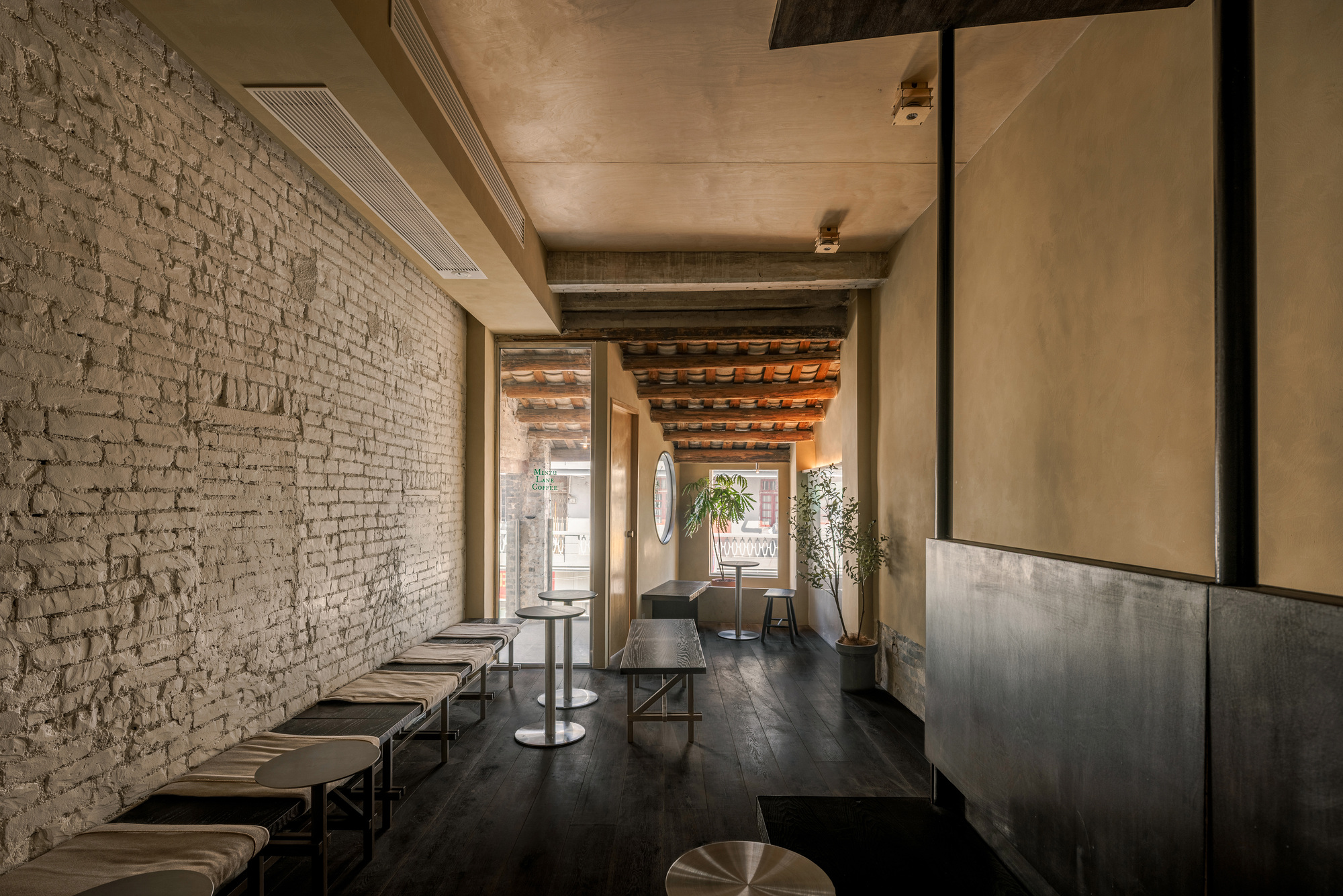 Narrow café room with whitewashed brick wall, low cushioned benches, dark timber floor and exposed terracotta-tiled rafters in soft daylight