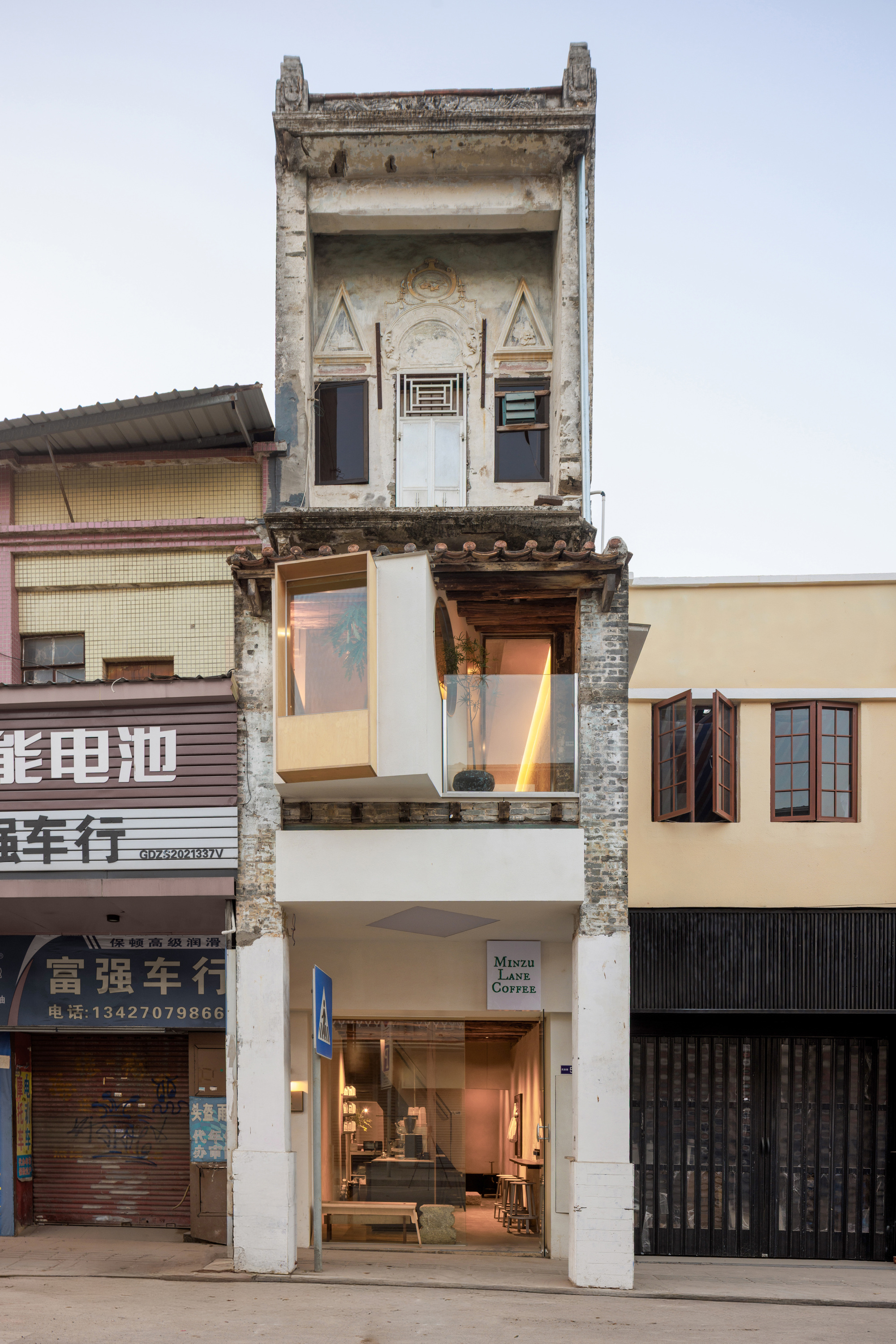 Street facade of a narrow weathered building with a glass and timber box projecting from the upper floor at dusk