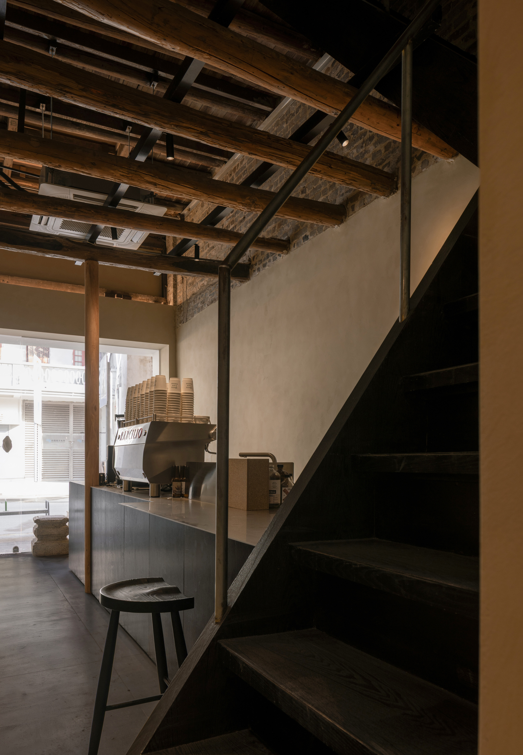 Coffee counter with espresso machine beside a dark timber staircase, under exposed log joists and a bright street window