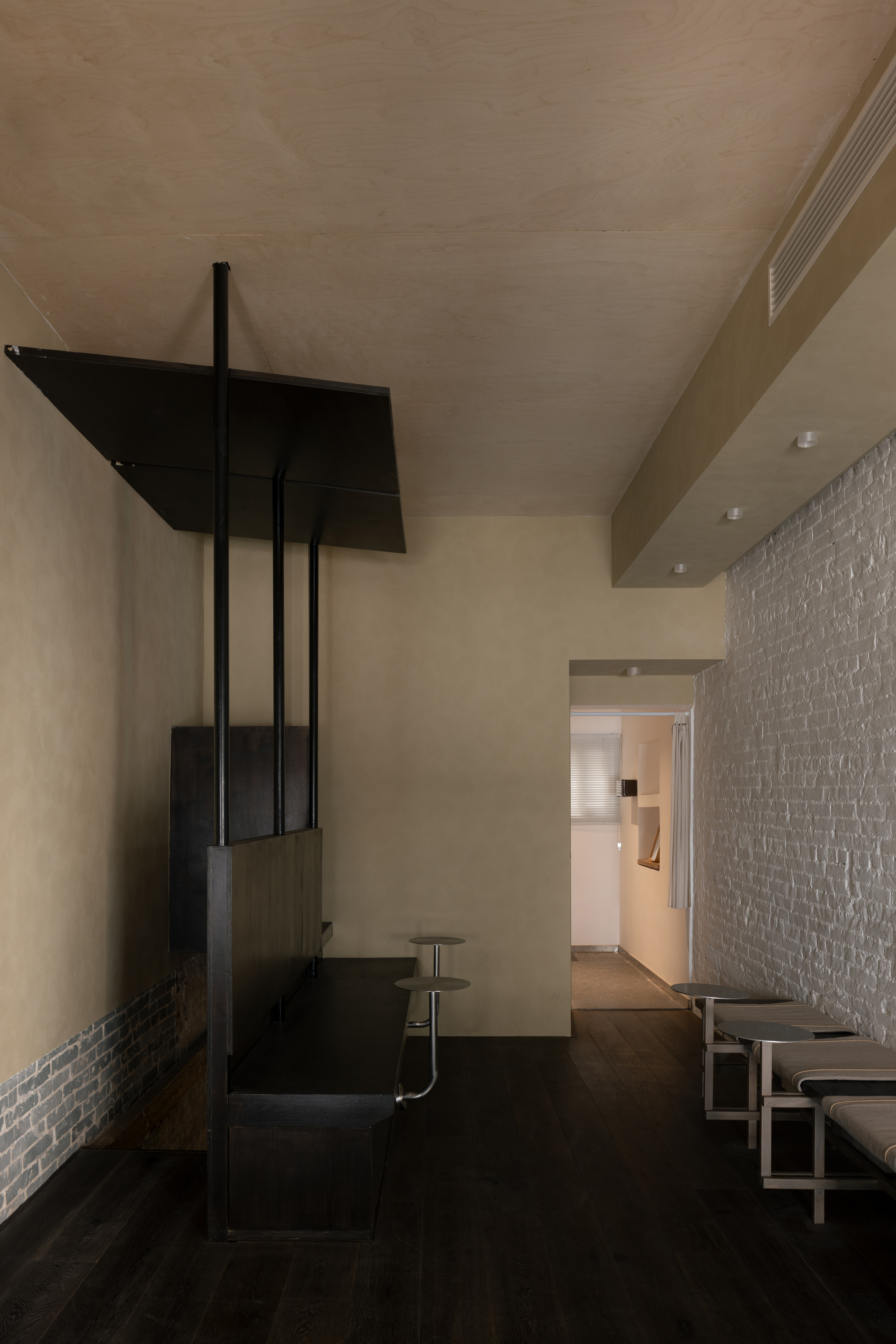Plaster-walled room with a dark suspended steel and timber canopy, grey brick wall and slim metal stools on a dark floor
