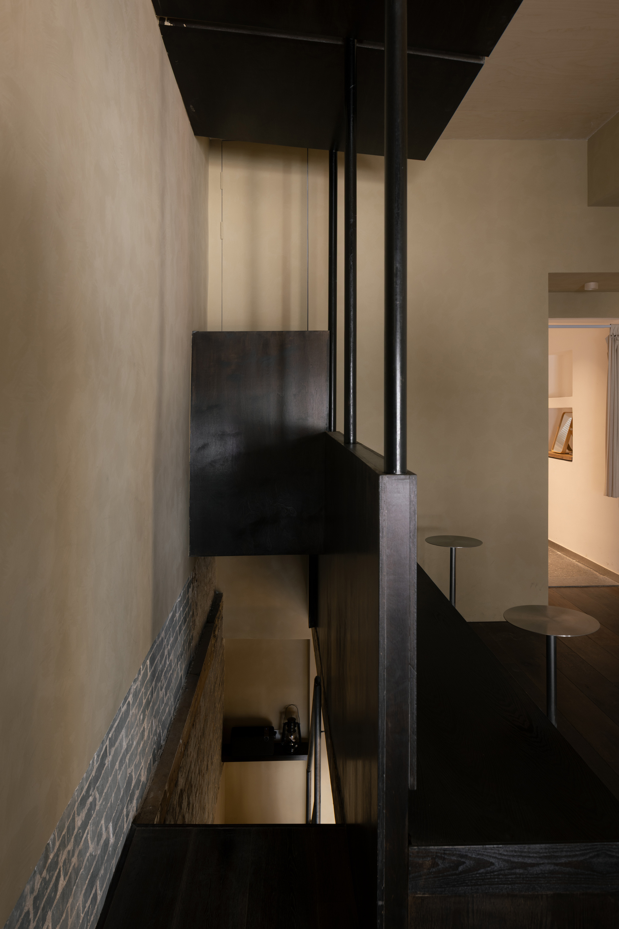 Looking down the stairwell past a dark timber balustrade and steel rods toward small round tables and a lit doorway
