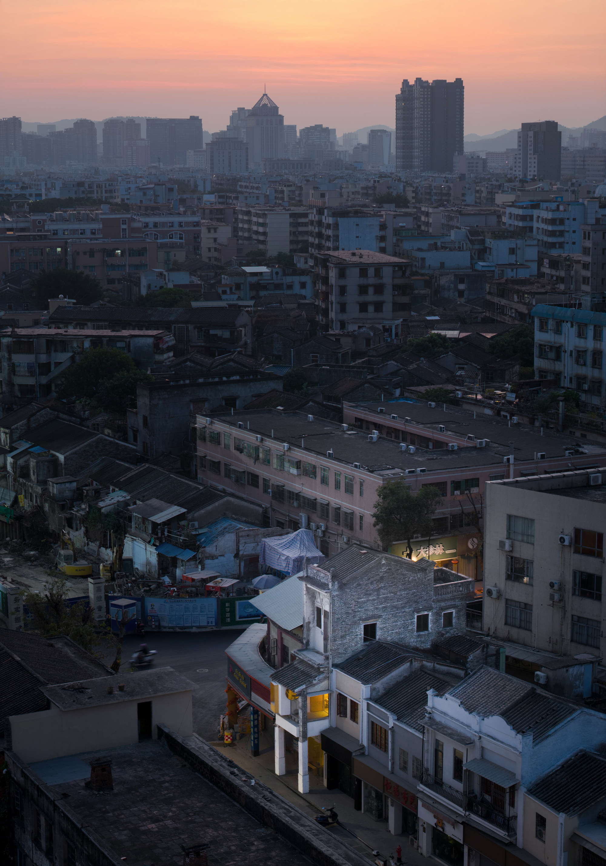 Elevated view over low rooftops and a street corner of small shopfronts toward distant towers under an orange dusk sky