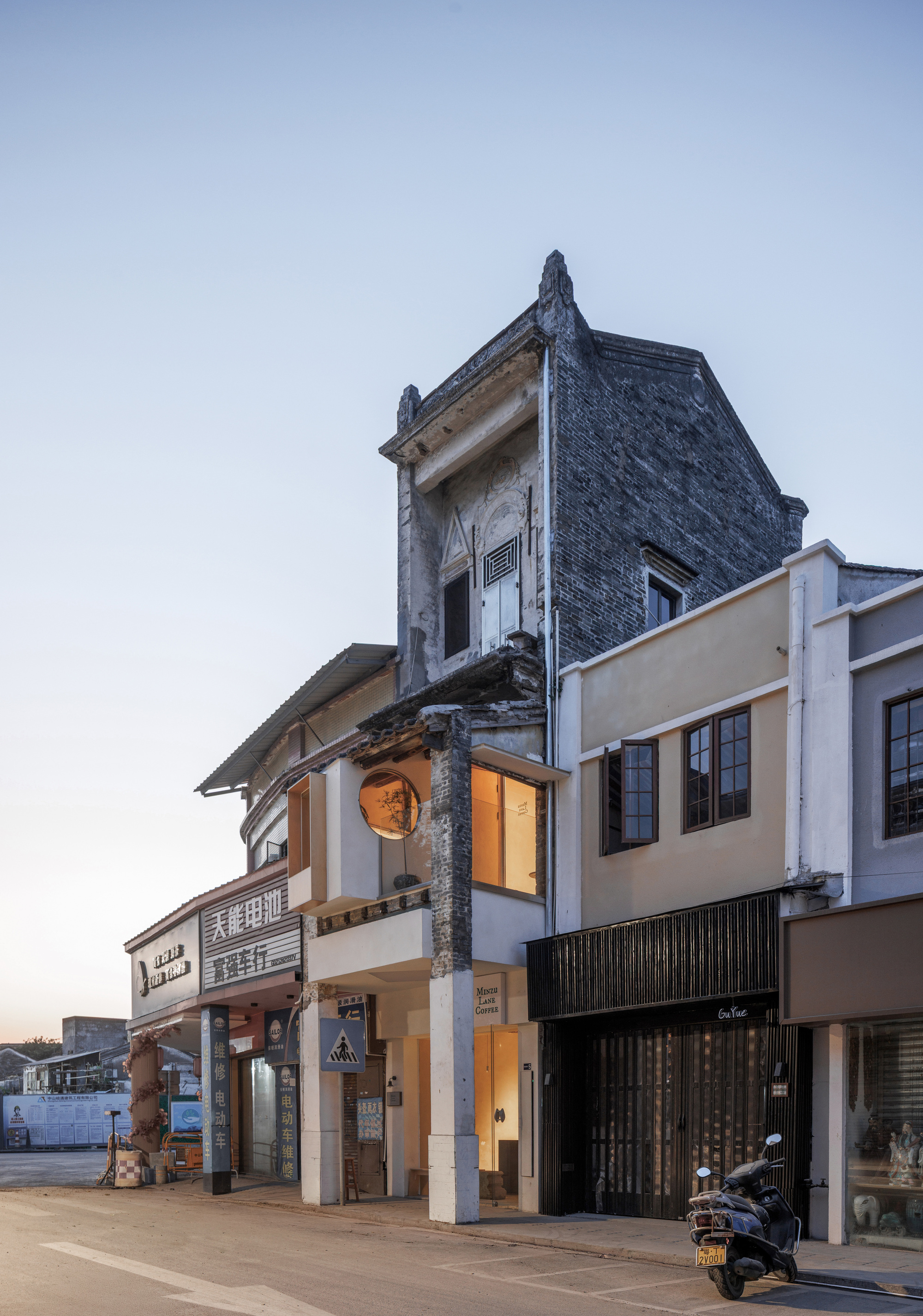 Street facade of a narrow building with a weathered grey brick gable, round window and a parked scooter at dusk