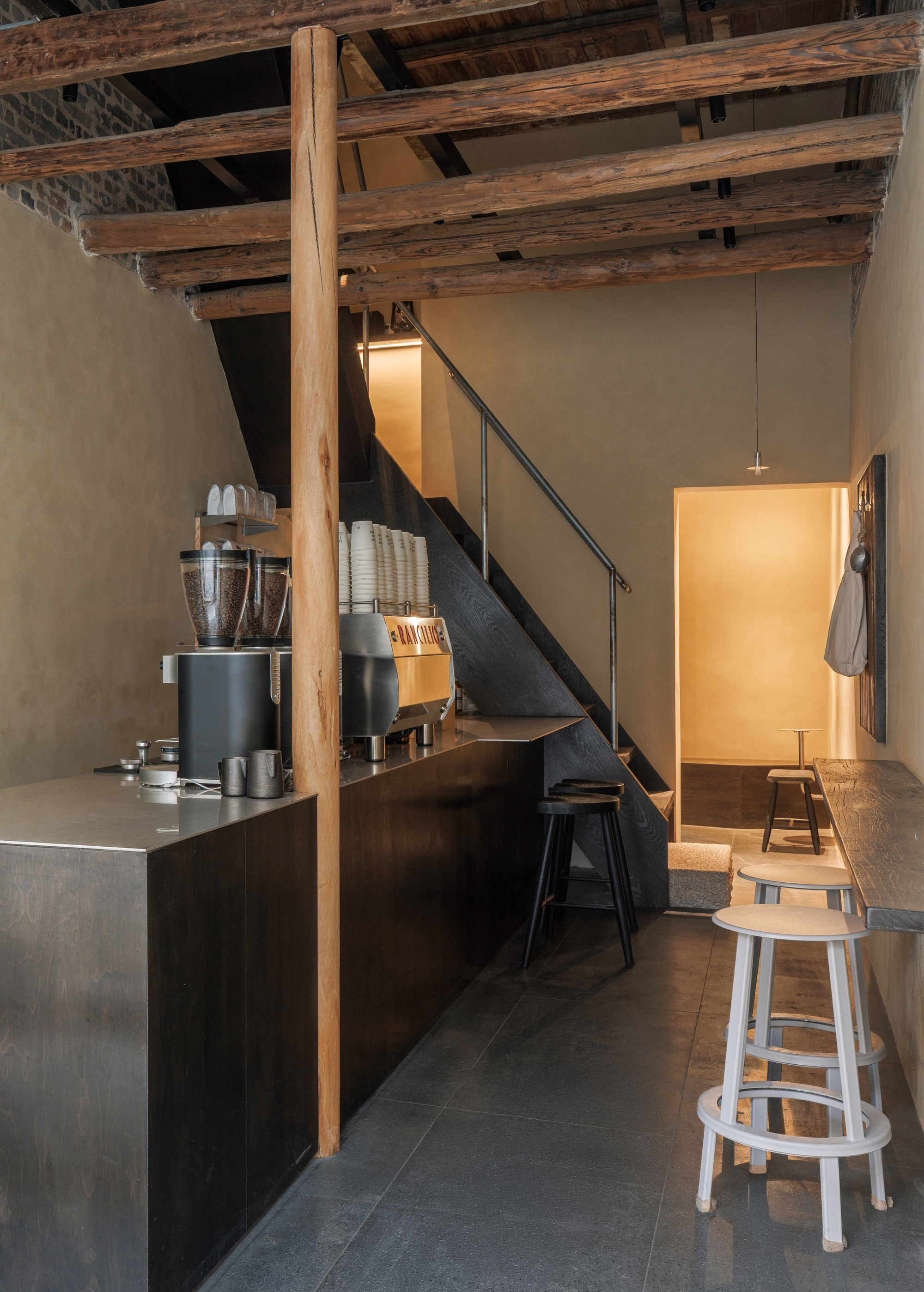 Coffee bar interior with a steel-topped counter, exposed timber rafters, a dark staircase and pale stools in warm light