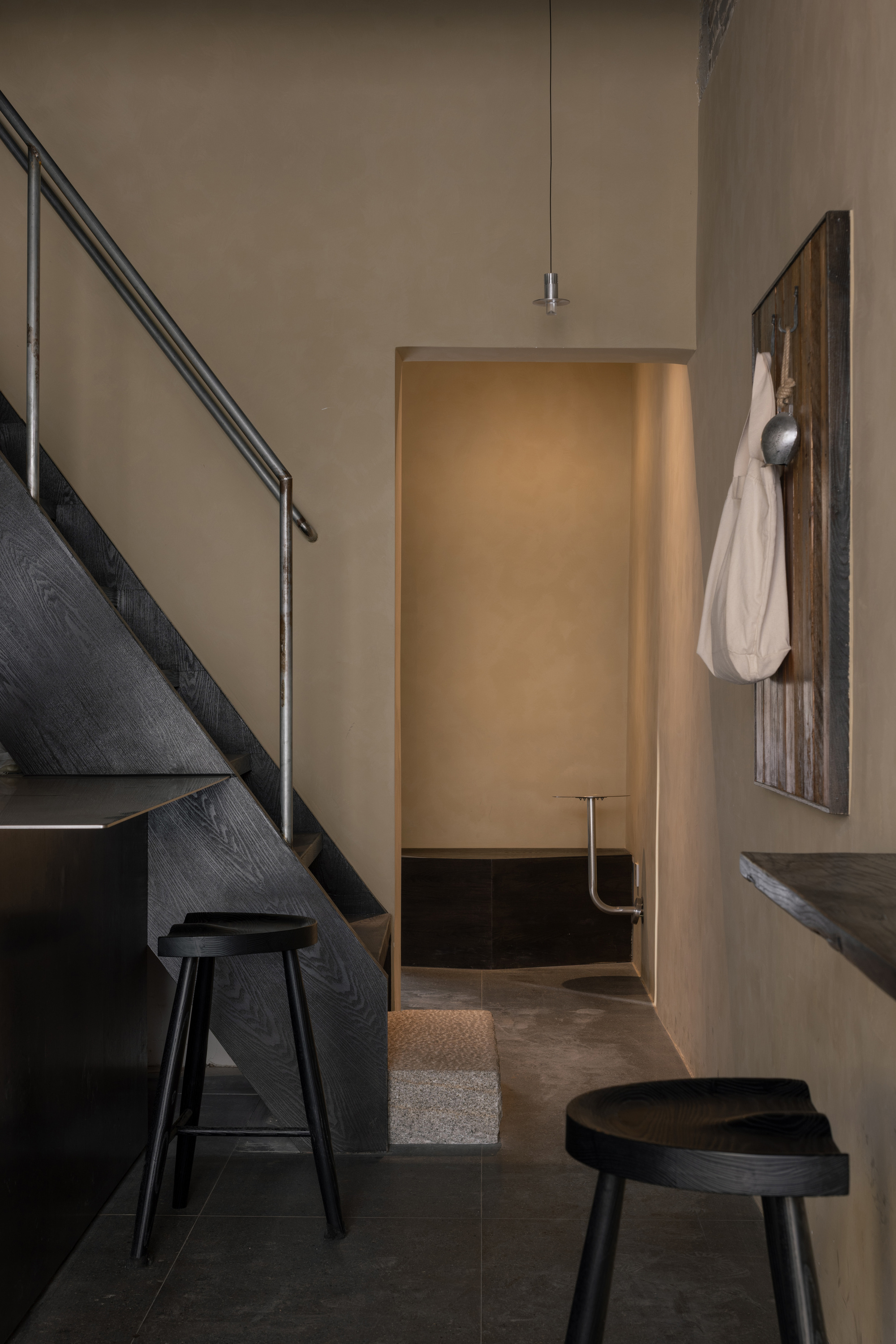 Corner of the cafe with a blackened timber staircase, steel handrail, black stools and a glowing doorway beyond