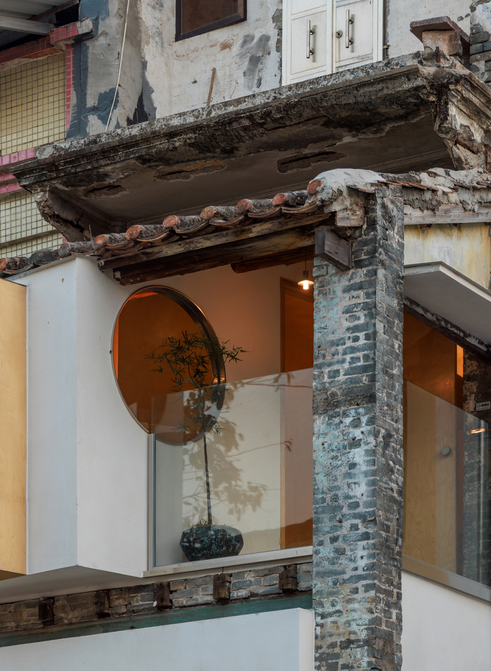 Close-up of the weathered brick column and clay tile eave beside a round window with a potted tree, lit at dusk