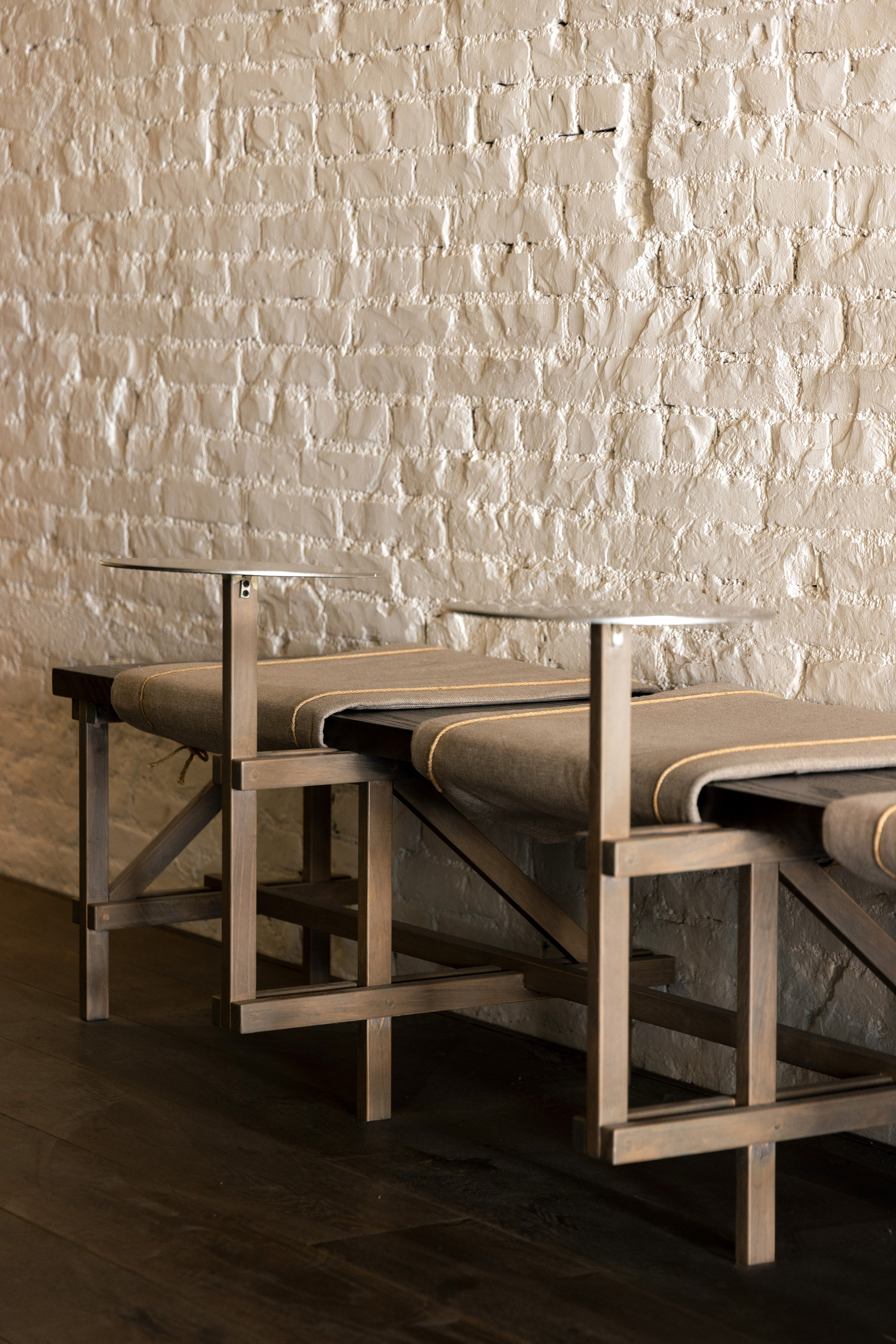 Pair of timber benches with canvas cushions and small metal shelves against a white-painted brick wall, over a dark plank floor