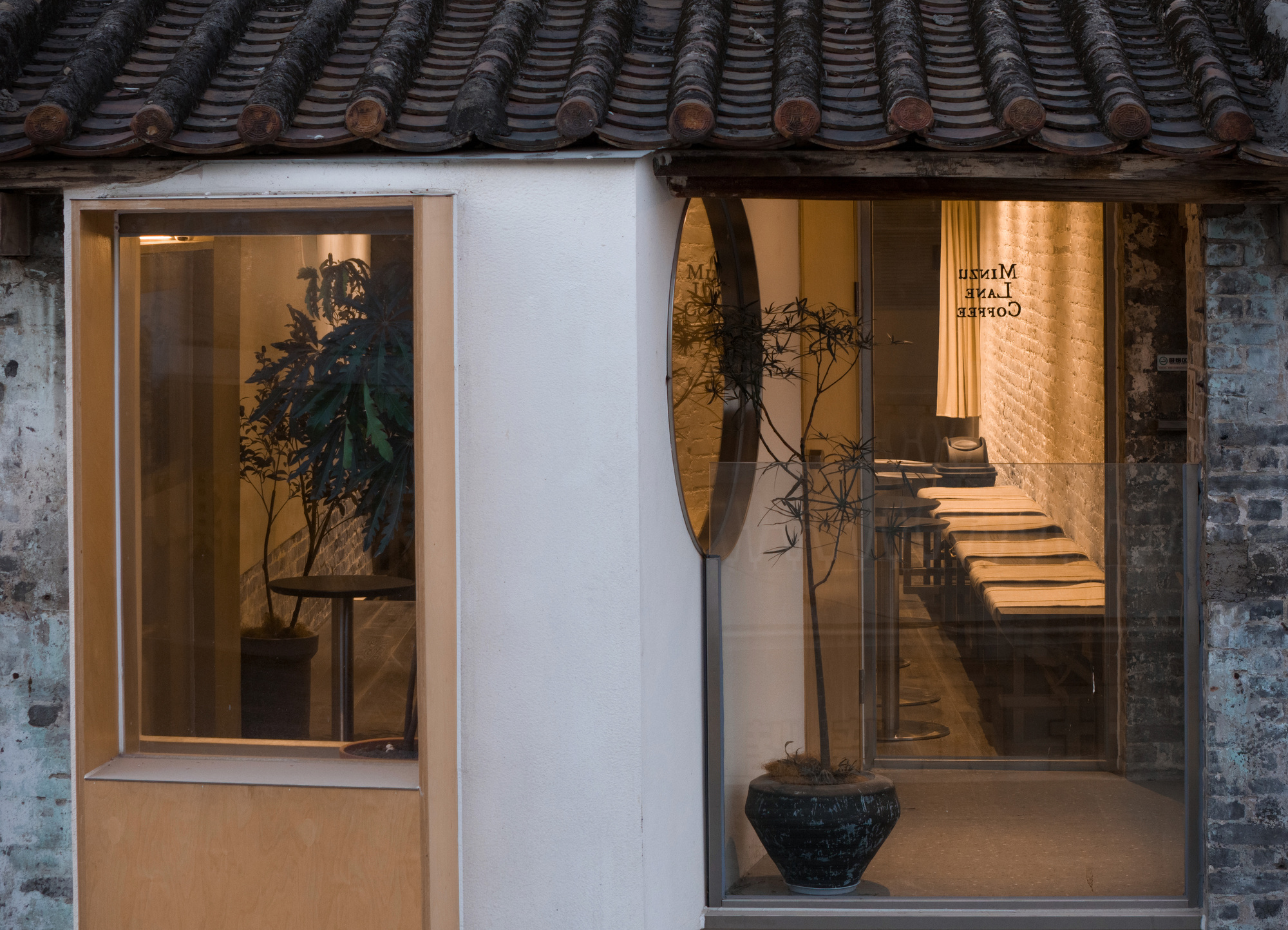 Shopfront with a plywood-framed window and glazed entrance, a potted tree inside, beneath an old tiled eave at dusk