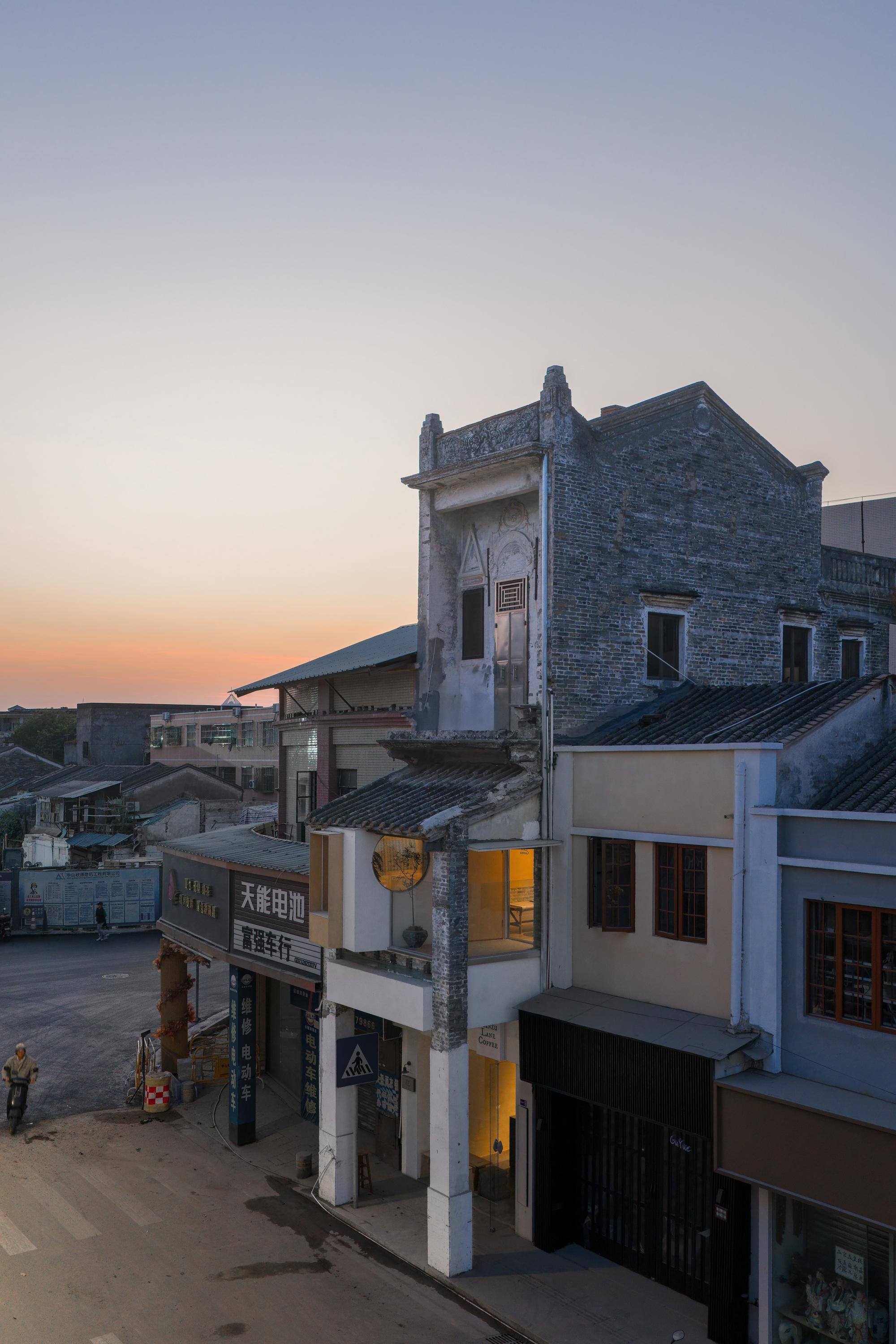 Elevated view of a narrow shopfront wedged between older brick houses on a street at sunset, a scooter rider below