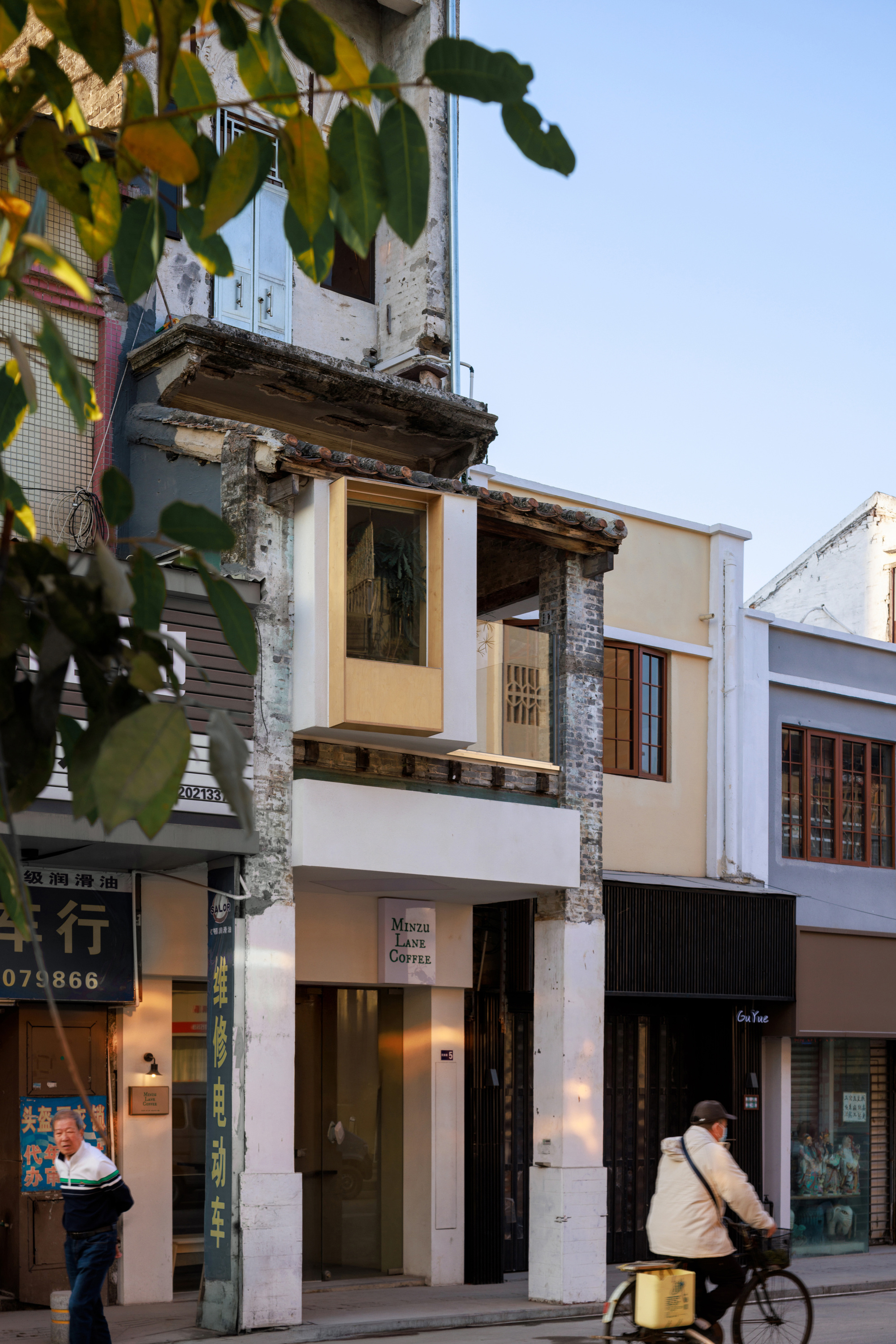 Street-level view of the narrow cafe facade with white columns and a projecting wood-framed window, a cyclist passing and leaves in the foreground