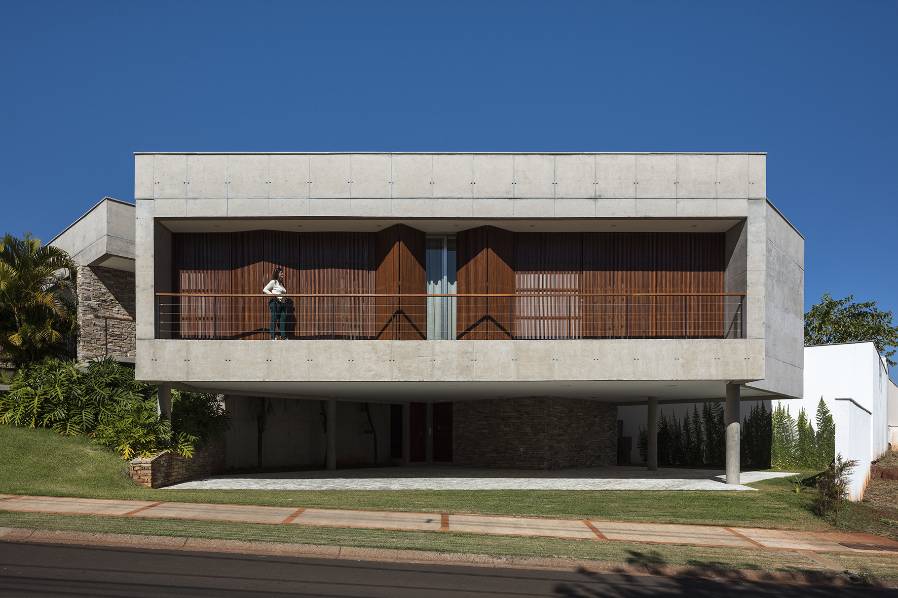 Street facade of a board-formed concrete box with a timber-screened balcony, a person at the railing, and a clear blue sky