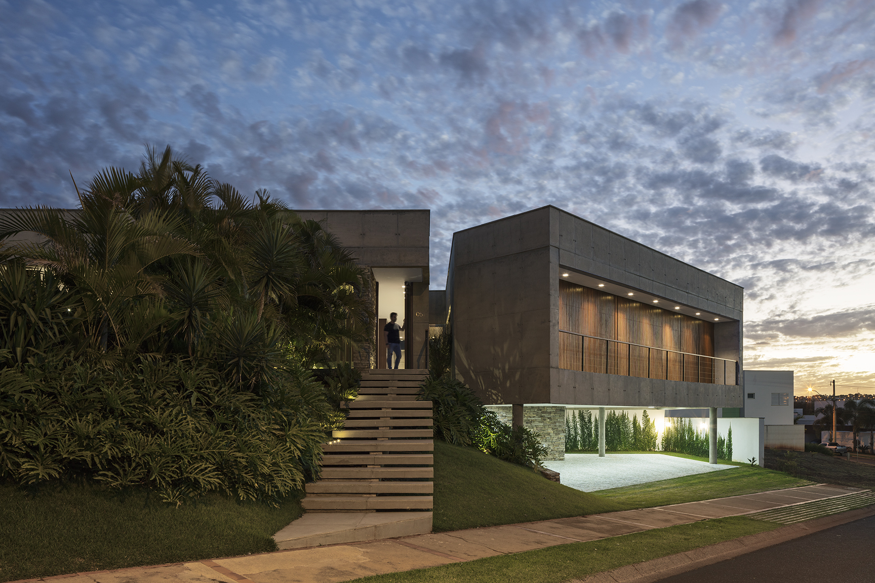 Floating concrete upper volume beside a slatted timber stair and dense palms, seen at dusk under a mottled cloudy sky