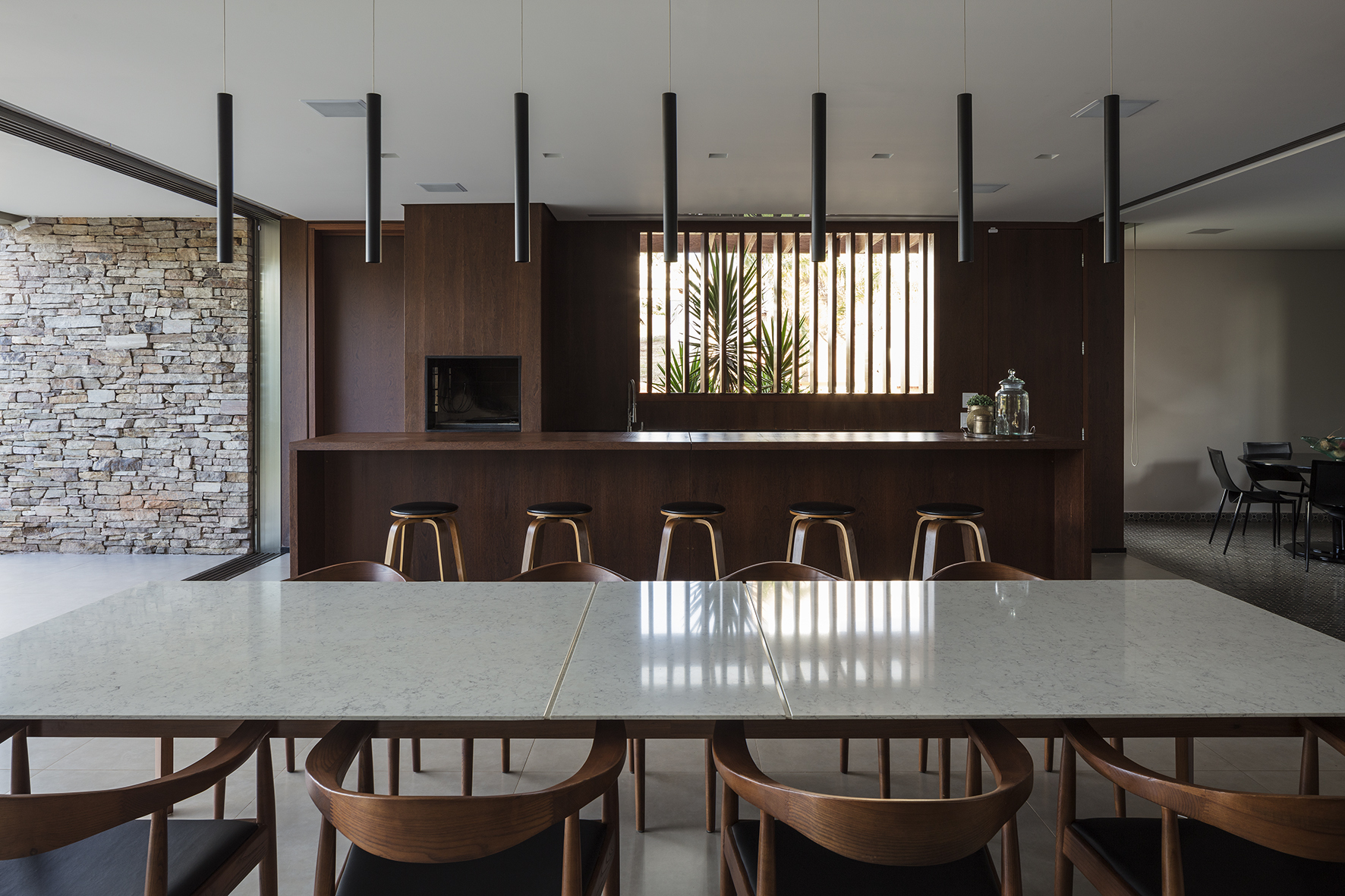Dining area with a stone-topped table, walnut chairs, a kitchen bar with stools, and black pendant lamps against a stacked stone wall
