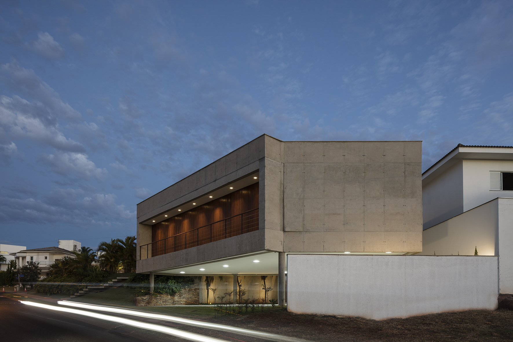 Side elevation of the raised concrete volume with lit timber-lined balcony and a white boundary wall, with car light trails on the road at dusk