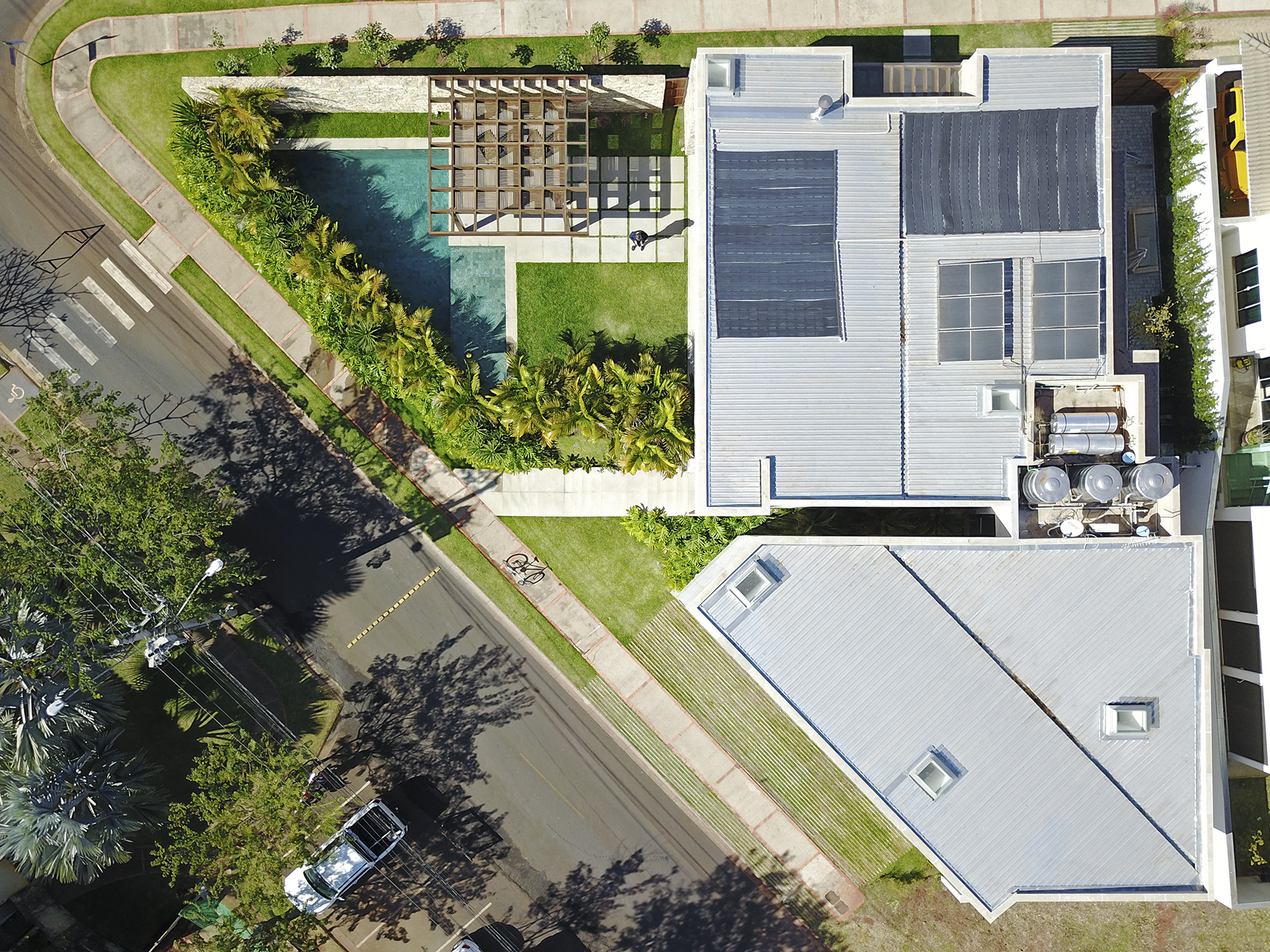 Aerial top-down view of metal roofs with solar panels and water tanks beside a pool, pergola and palm planting along the street