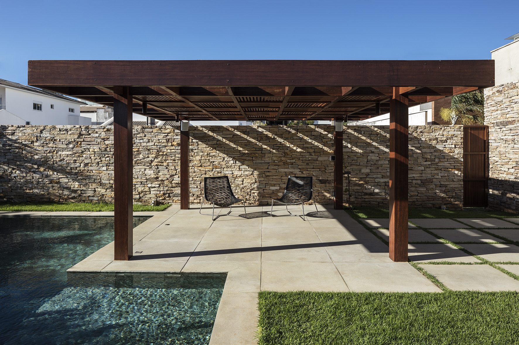 Timber pergola with dark wood columns over a paved terrace, two woven chairs, a stacked stone wall and pool edge under blue sky