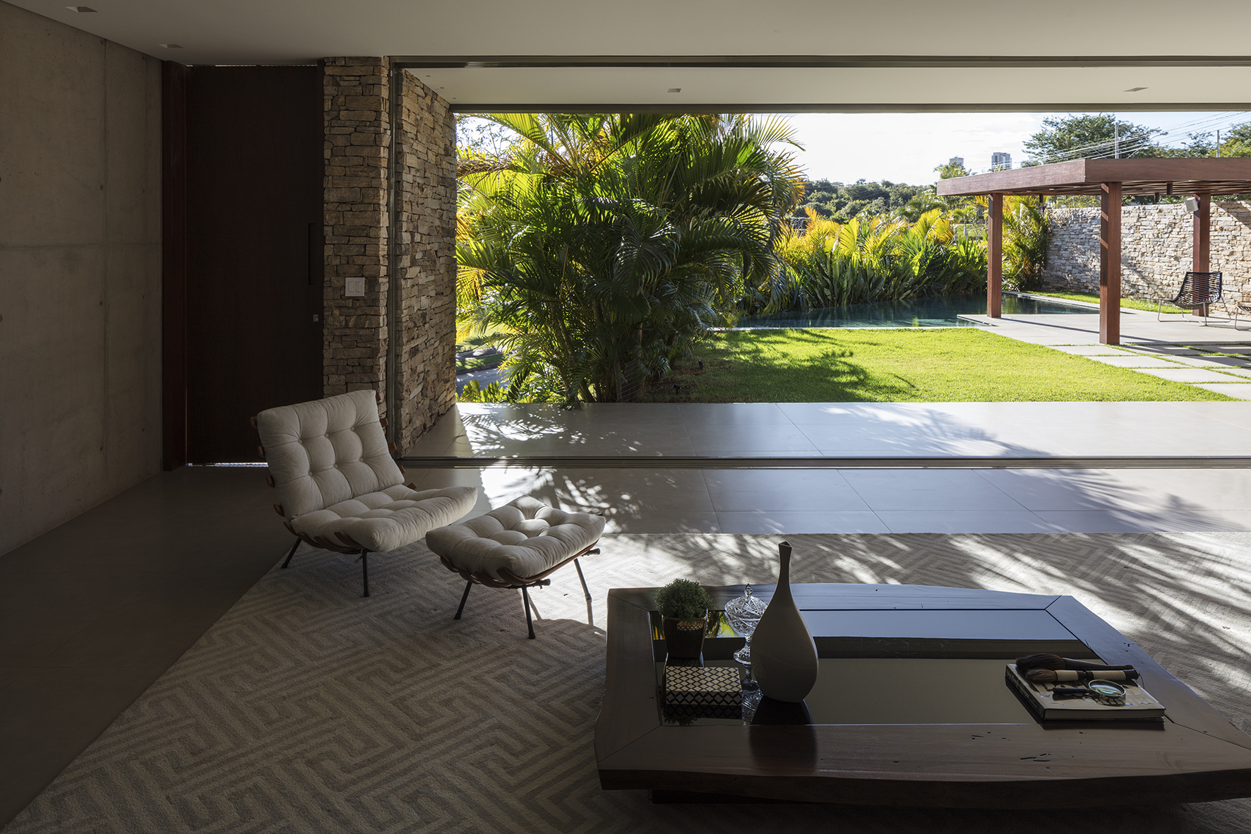 Living room with tufted lounge chair and ottoman, low wooden table and a sliding glass wall opening to palms and lawn in dappled sunlight