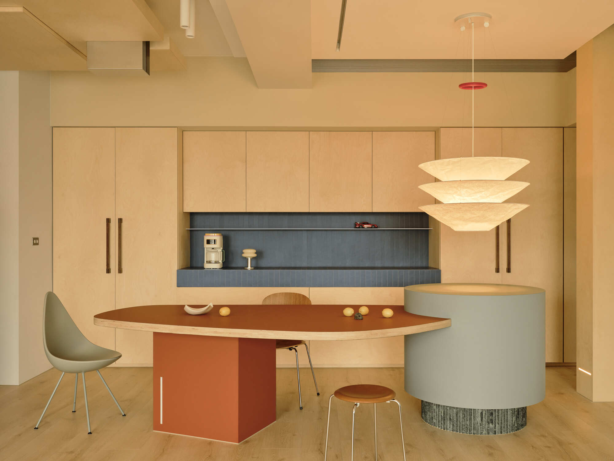 Plywood kitchen wall with blue tile backsplash, a leaf-shaped rust table and a tiered paper pendant glowing in warm light