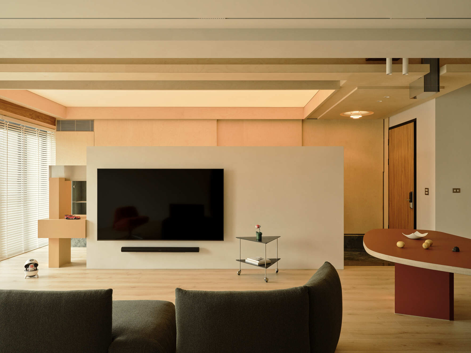 Freestanding white wall holding a wall-mounted television, with a gray sofa in the foreground and soft daylight through blinds